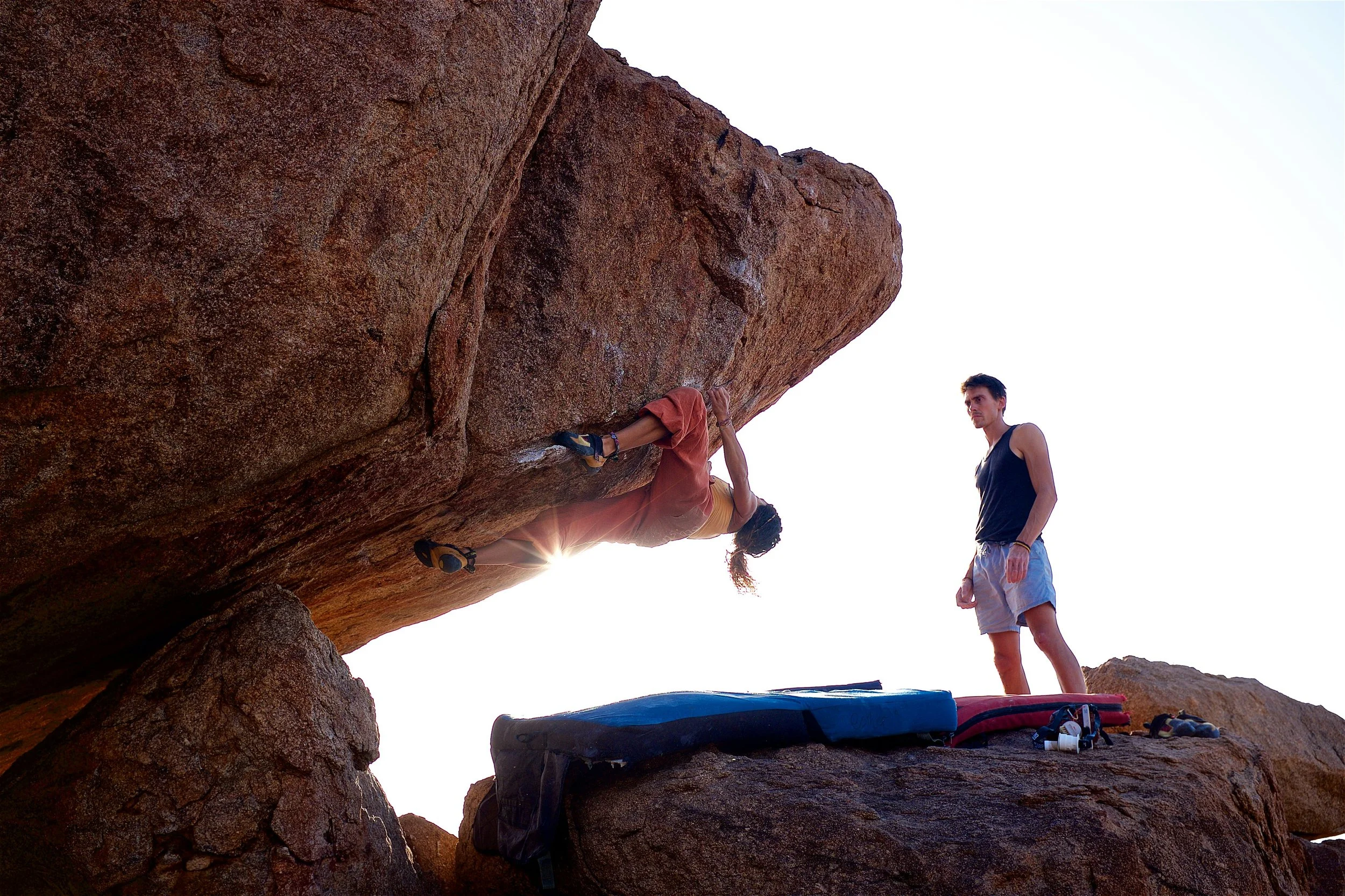 A woman rock climbing on a large, overhanging rock formation with a man standing nearby on a boulder, with climbing gear and crash pads on the ground.