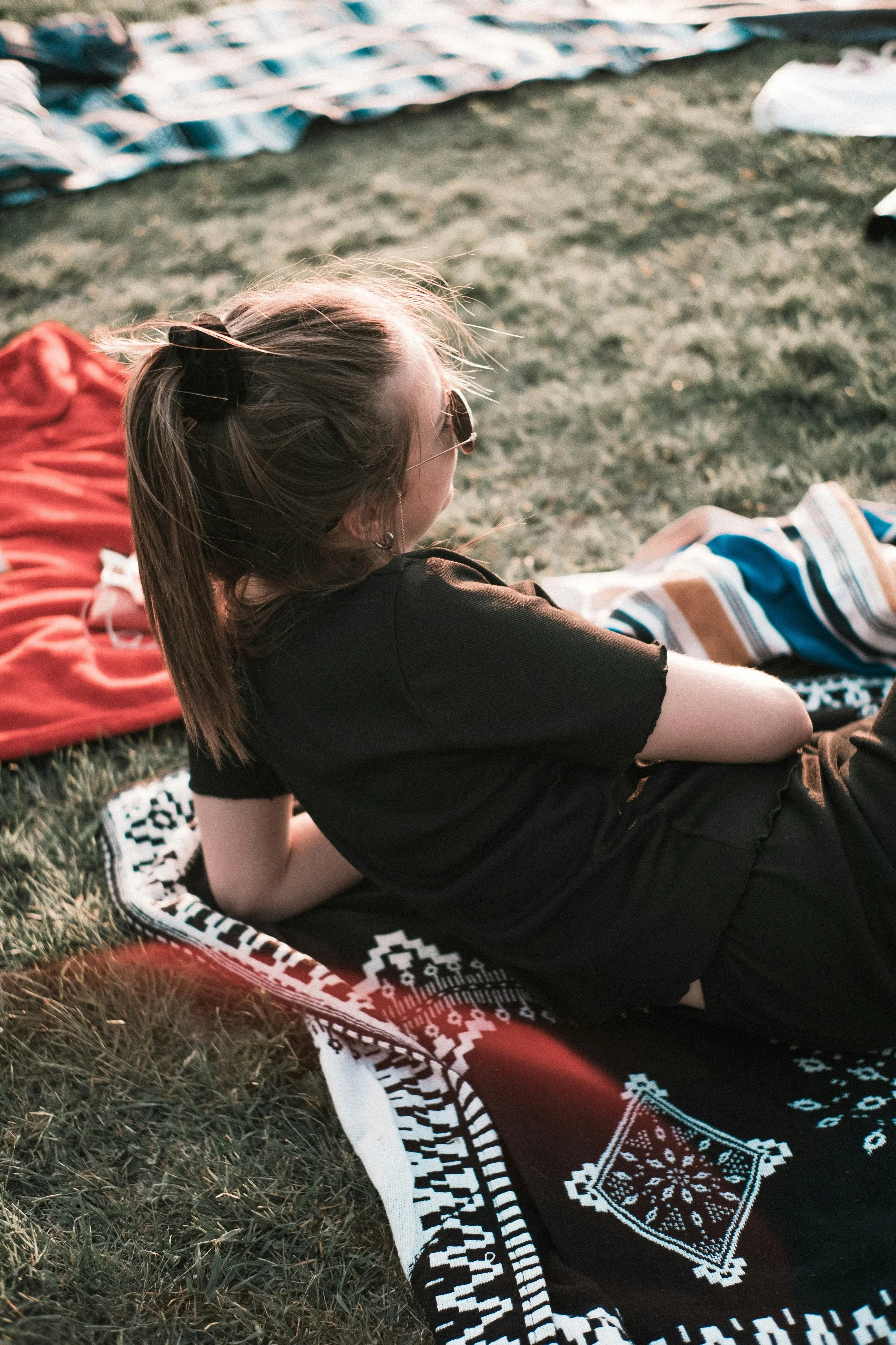 A girl with blonde hair tied in a ponytail, wearing sunglasses and a black t-shirt, is lying on her side on a patterned blanket on grass, looking away from the camera. There are other blankets and clothing items on the grass nearby.