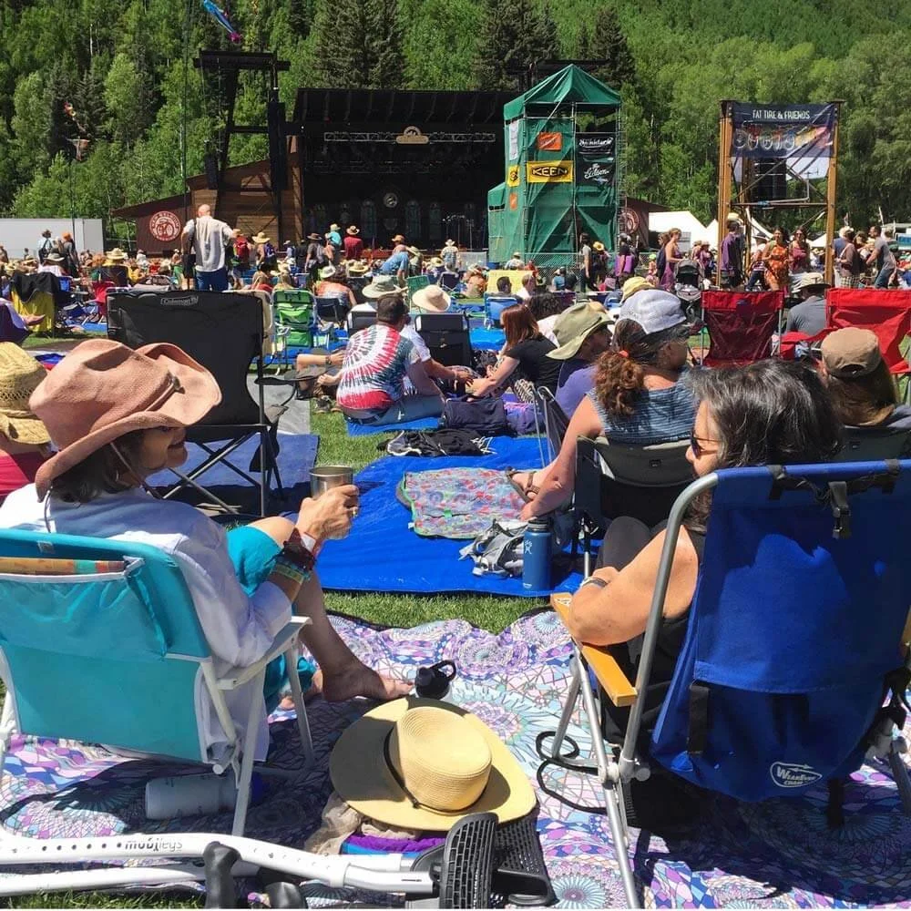An outdoor music festival with a large audience sitting on blankets and lawn chairs facing a stage set against a backdrop of green trees and hills. Performers are on stage with a screen and banners.