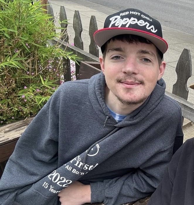 A young man with brown hair and a slight beard sitting outdoors on a wooden bench, wearing a dark gray hoodie and a black and red baseball cap that says 'Red Hot Chili Peppers,' next to plant greenery and a wooden fence.
