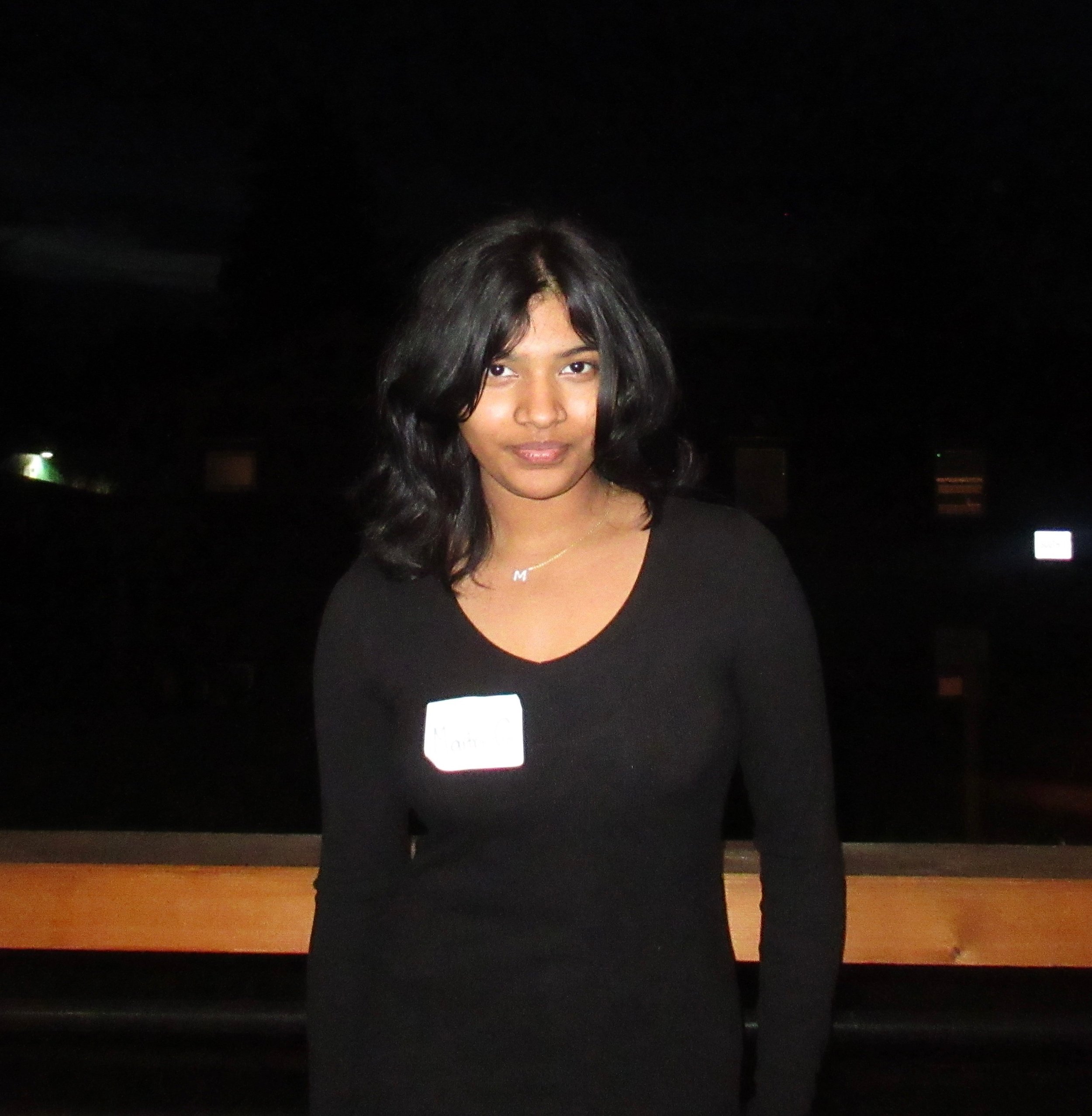 A young woman with shoulder-length black hair, wearing a black long-sleeve top, stands at night against a dark background with some distant lights, and has a name tag on her chest.