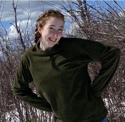 Young woman smiling outdoors during winter, standing in front of bare trees and with a cloudy sky.