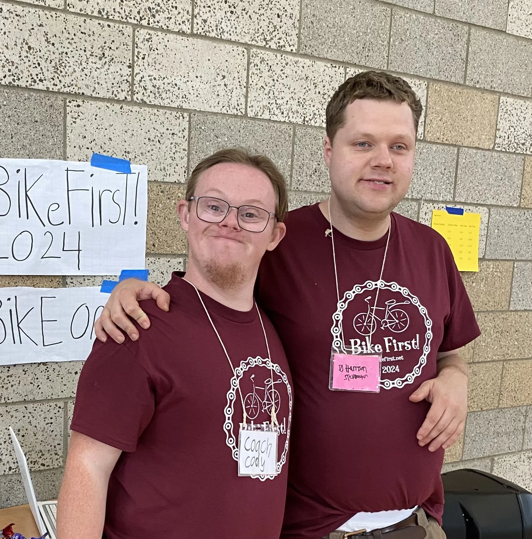 Two men wearing maroon T-shirts with 'Bike First!' logo, standing arm-in-arm indoors against a brick wall with signs about a bike event.