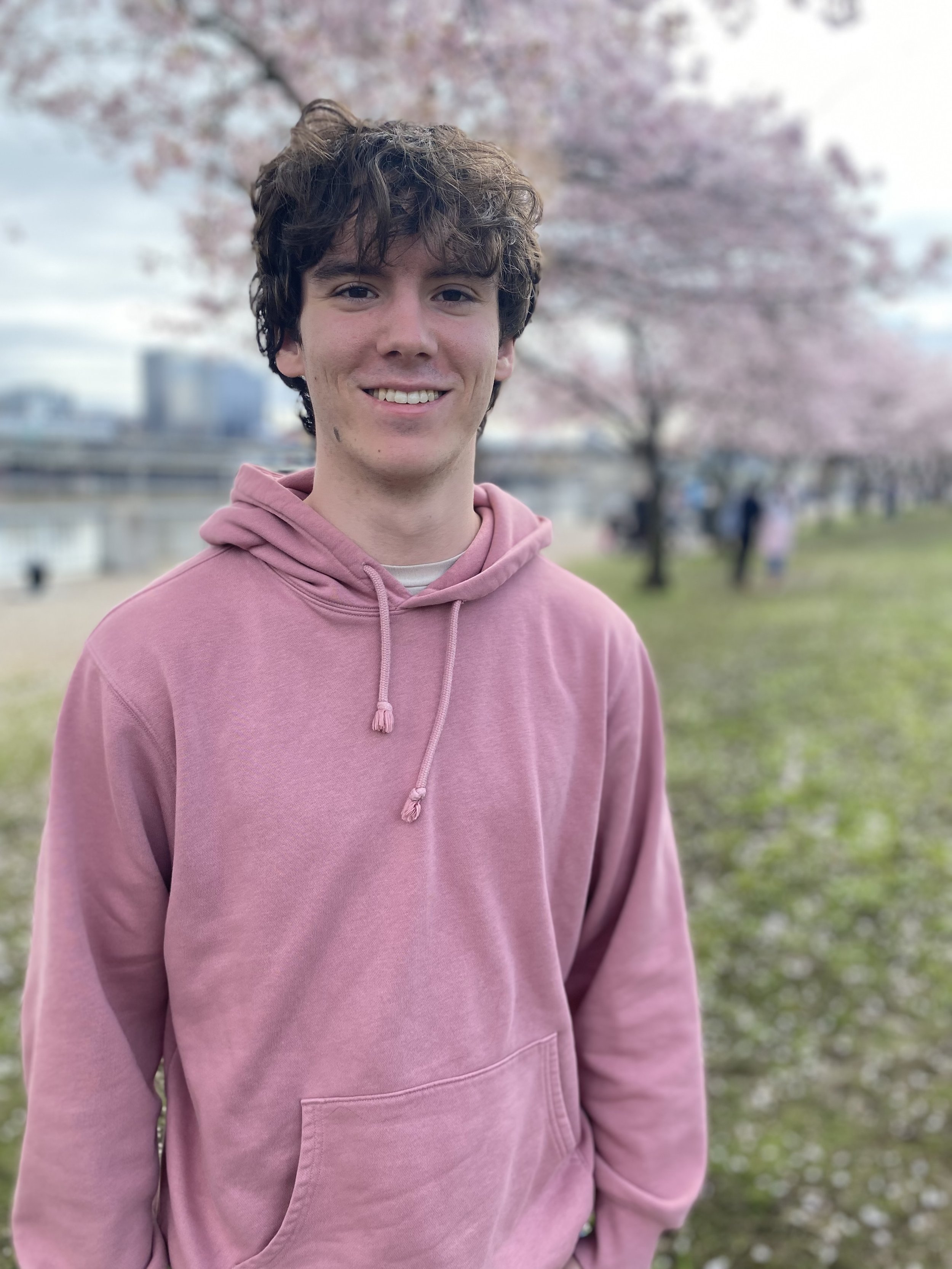 A young man with curly brown hair and a pink hoodie smiling outdoors near cherry blossom trees with a cityscape in the background.