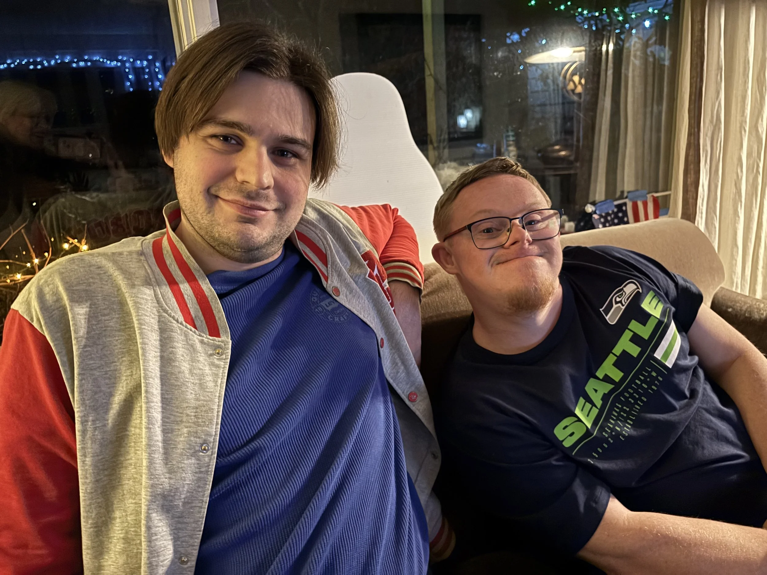 Two men sitting on a couch in a cozy room with holiday decorations, smiling at the camera.