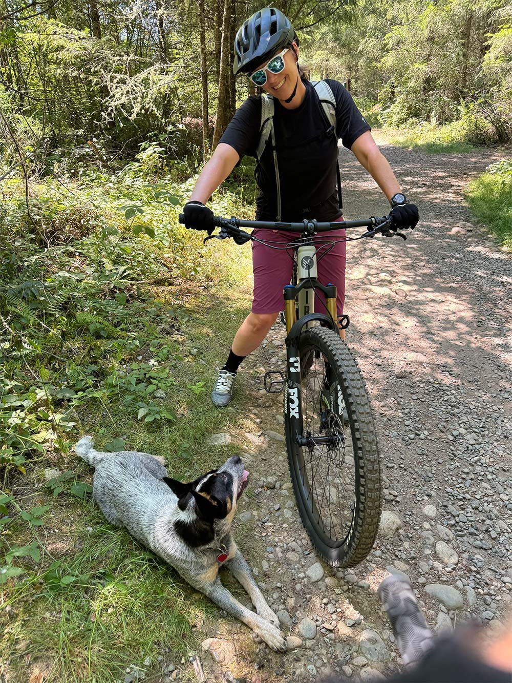 Kristen mountain biking through a forest trail with her dog, showing balance, energy, and playful leadership.
