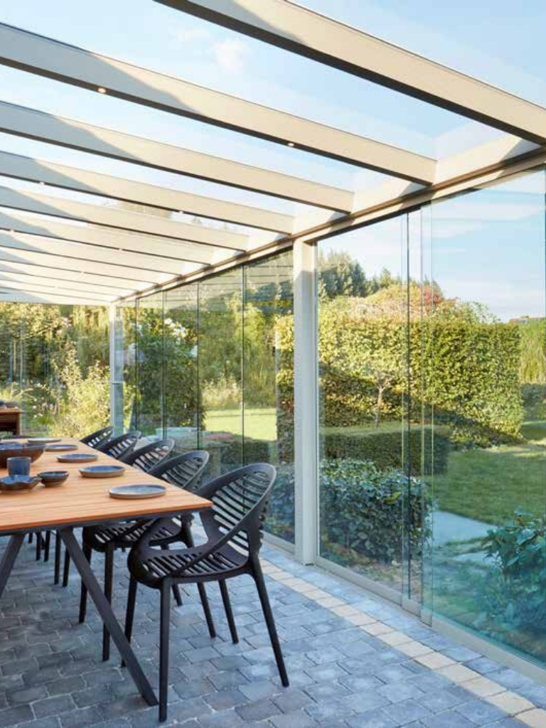 Terrasse avec pergola en aluminium, toiture en verre, avec une table en bois et des chaises noires, surplombant un jardin verdoyant.