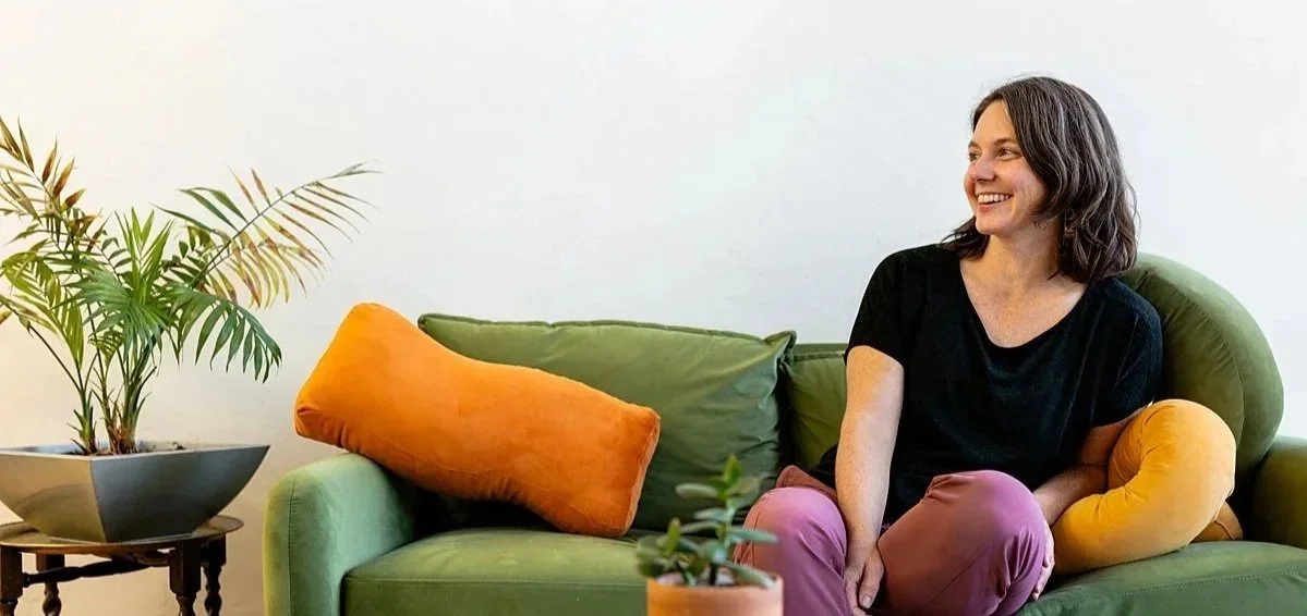 Katy, your physical therapist and coach, with shoulder-length dark hair sitting on a green sofa, smiling, with orange and yellow pillows, next to a potted plant in a gray and black pot, against a white wall.