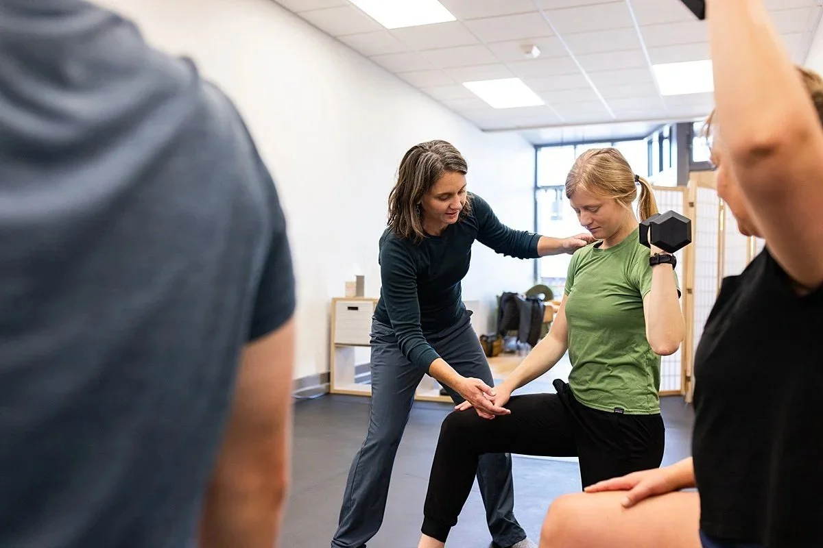 A woman providing physical therapy or exercise instruction to a young woman lifting a dumbbell in a rehabilitation or gym setting, with other individuals observing and practicing.