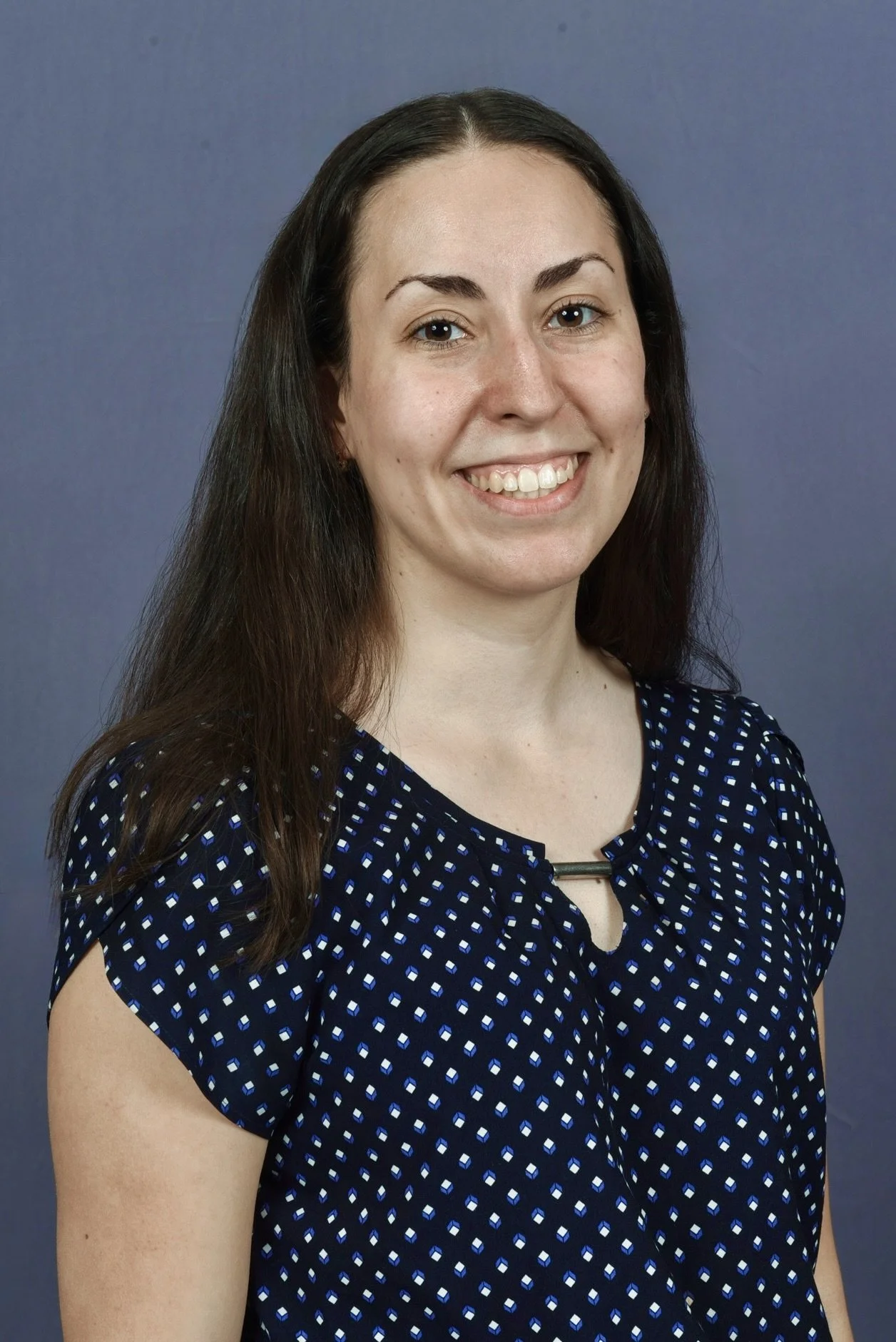 This is an image of Ashley Rodrigues, Research Assistant, and MA School and Applied Child Psychology student. A woman with long dark hair smiling, wearing a navy blue top, standing in a hallway with blurred background.