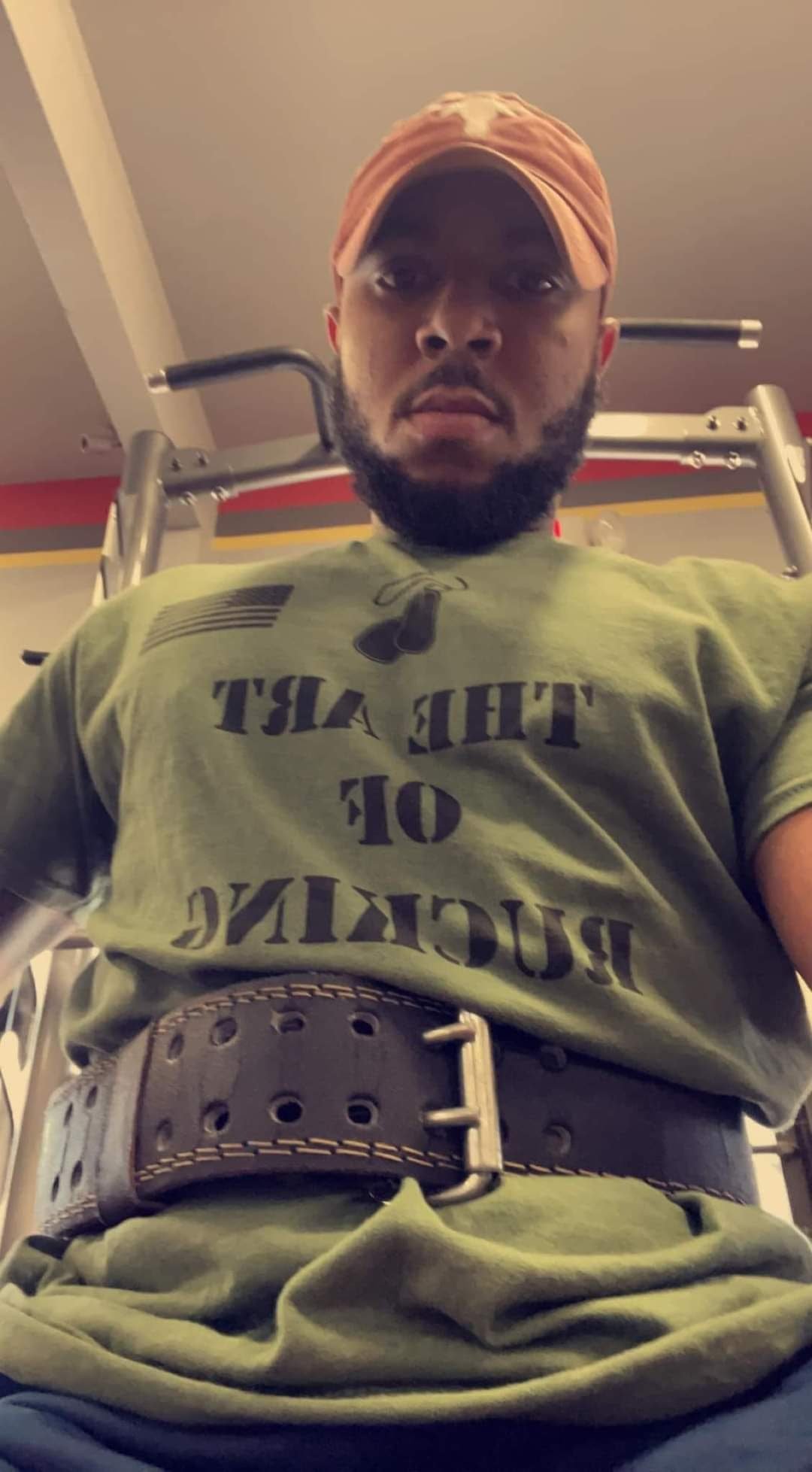 Man wearing a baseball cap and a green T-shirt in a gym, using a weightlifting belt.