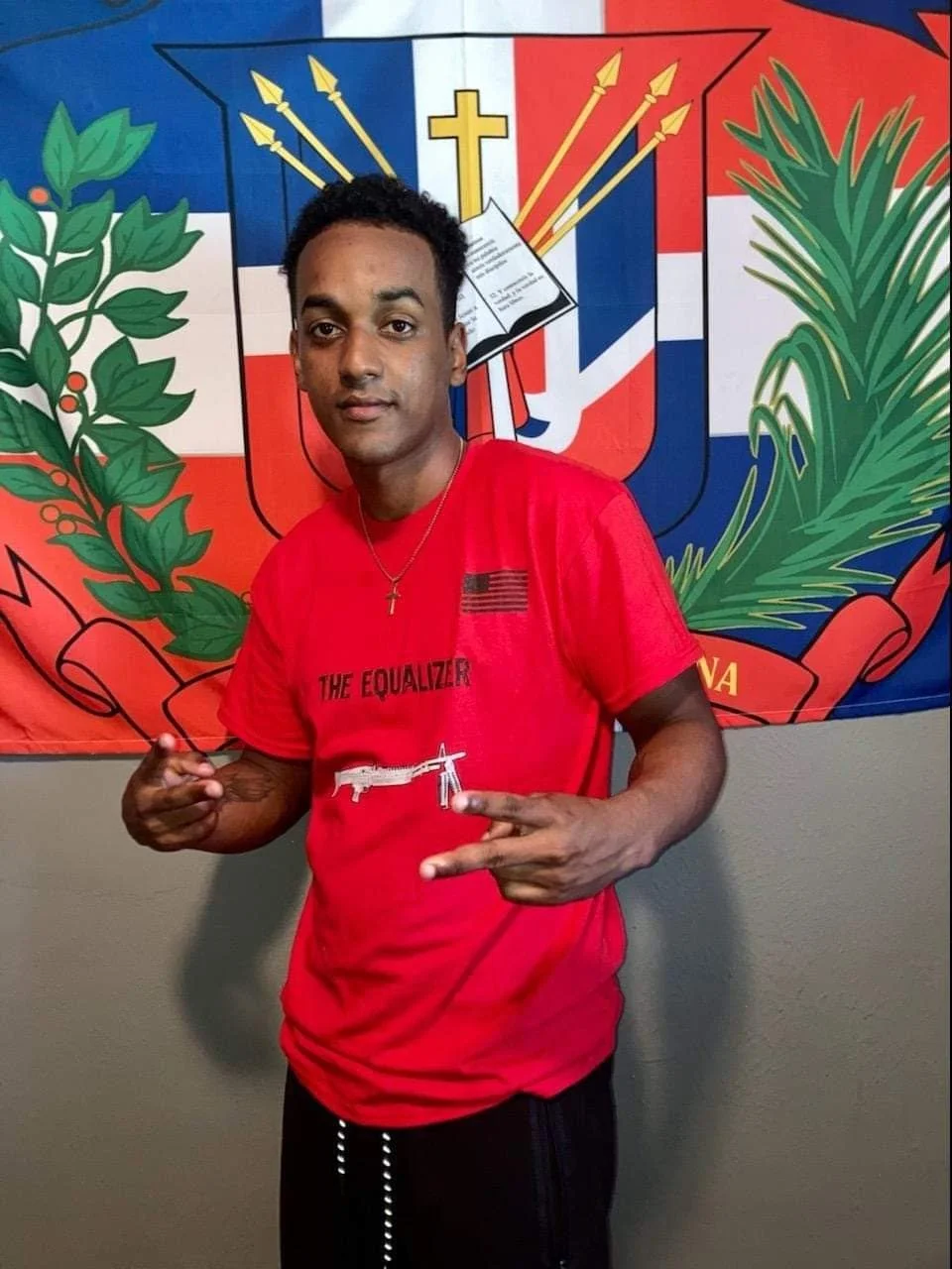 Person in red shirt with "The Equalizer" text, standing in front of a colorful flag or emblem background, making a hand gesture.