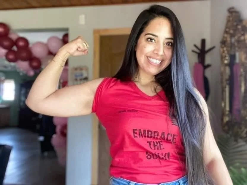 Smiling woman flexing arm in red shirt with "Embrace The Suck" text, indoors with balloons in background.