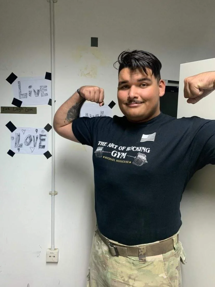 A person smiling and flexing their arms, wearing a black T-shirt and camouflage pants, standing in front of a wall with motivational signs taped to it.