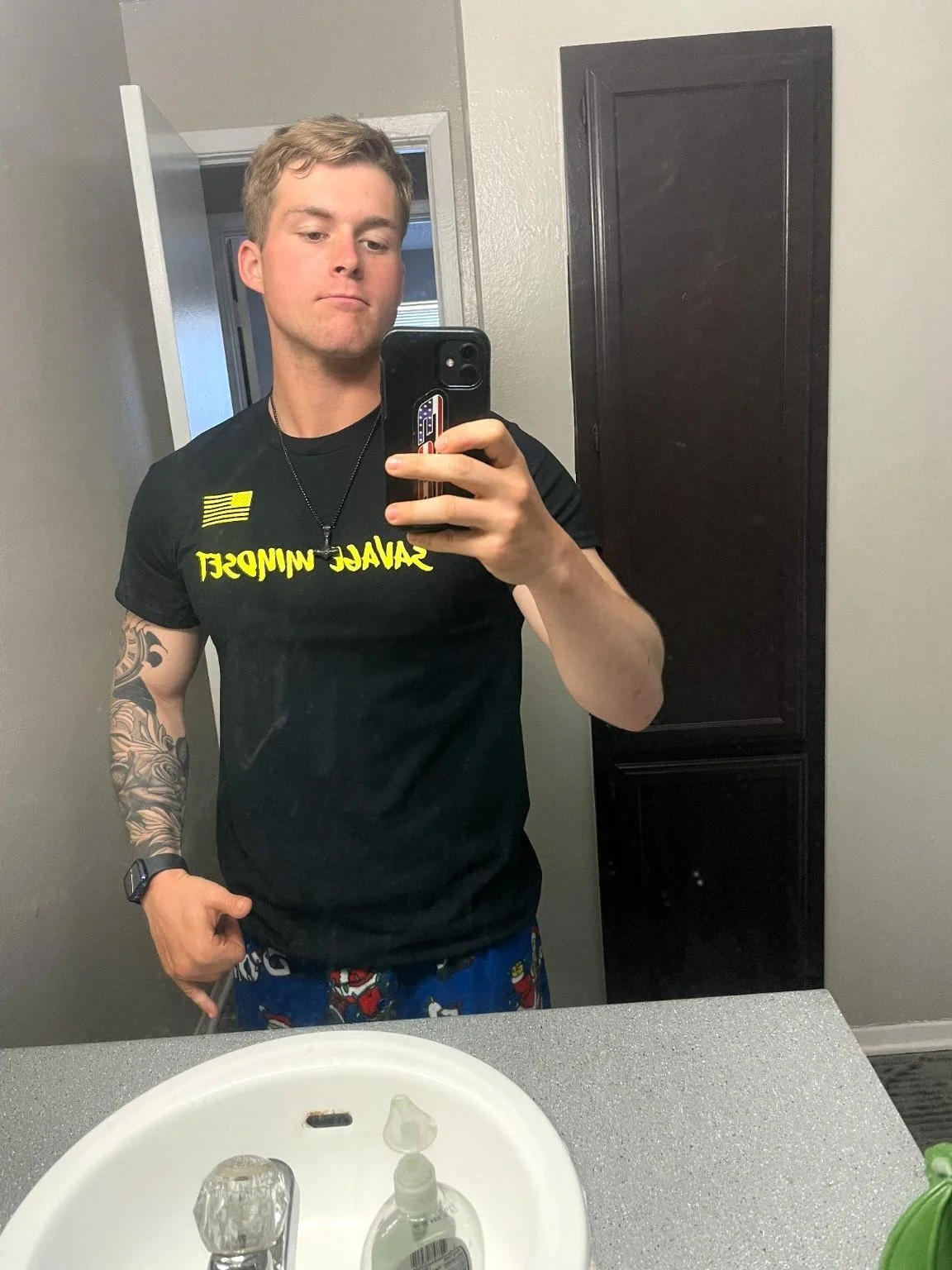 Person taking a selfie in a bathroom mirror, wearing a black T-shirt with yellow text and character-themed pajama pants, with a smartphone, and showing a tattooed arm.