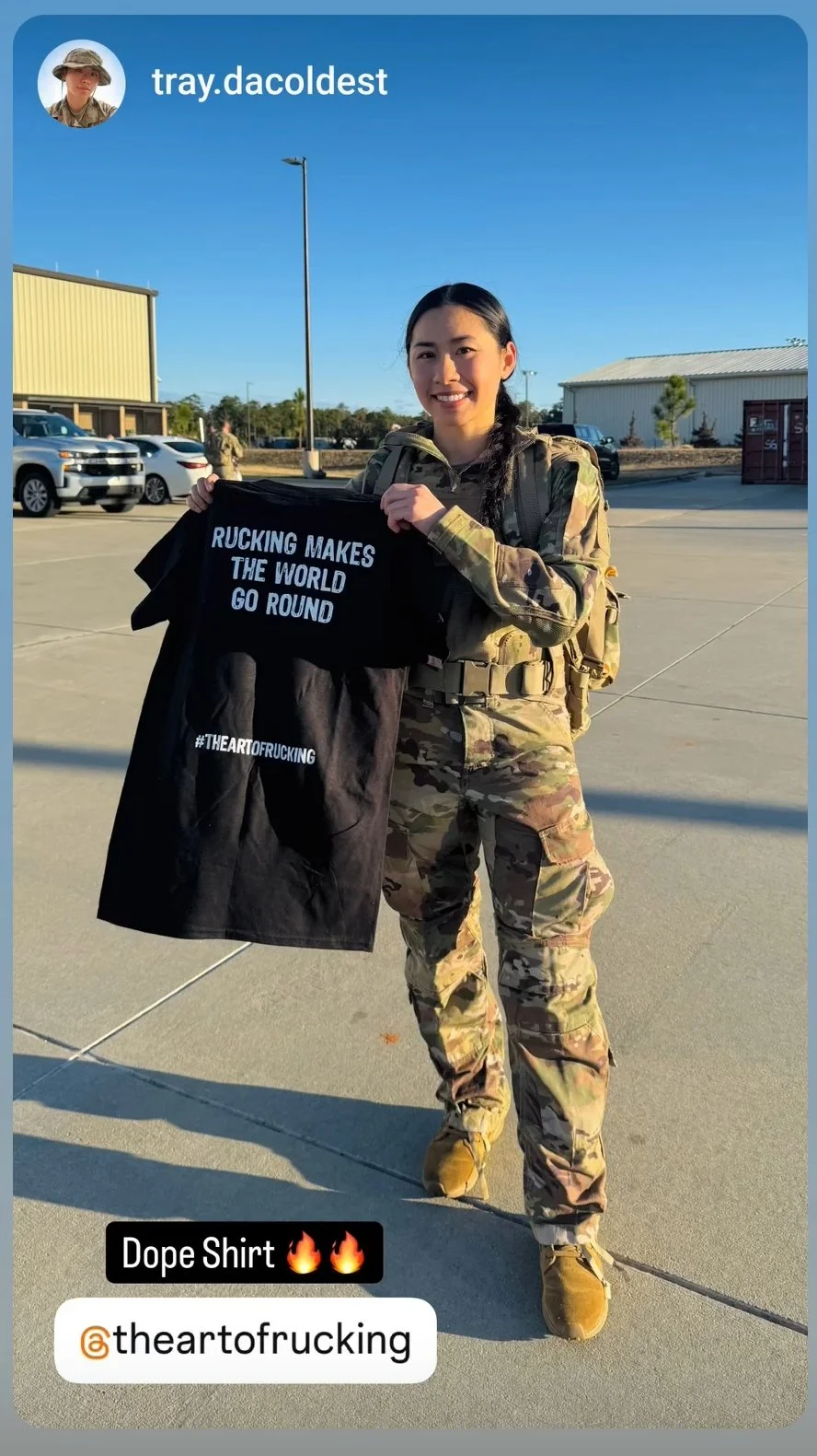 Person in military uniform holding a black T-shirt with the text "Rucking Makes the World Go Round" printed on it, outdoors with buildings and vehicles in the background.
