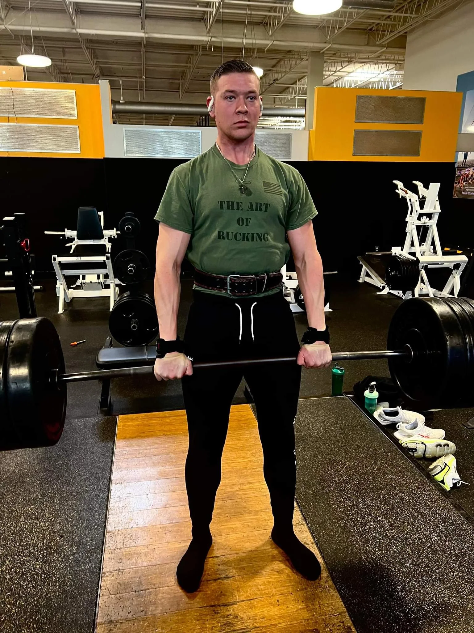 Person lifting a barbell in a gym, wearing a green T-shirt and black pants, with gym equipment in the background.