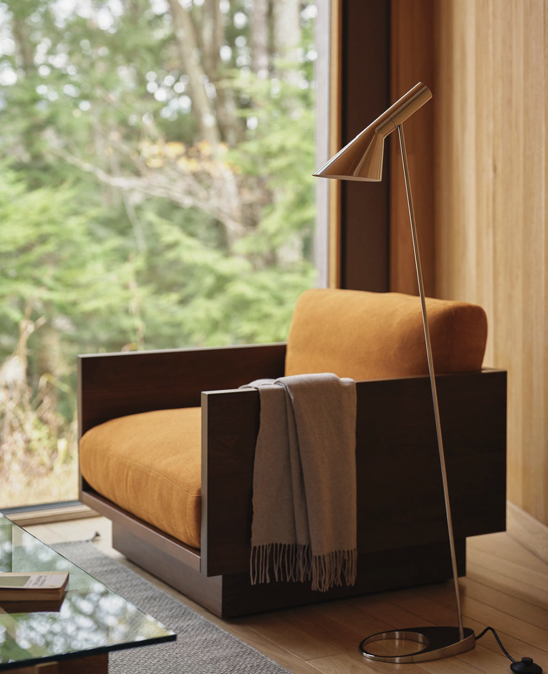 A modern armchair with orange upholstery and dark wooden sides, placed by a window overlooking a wooded area. A gray throw blanket hangs over the armrest, and a sleek floor lamp with a metallic finish stands next to the chair.