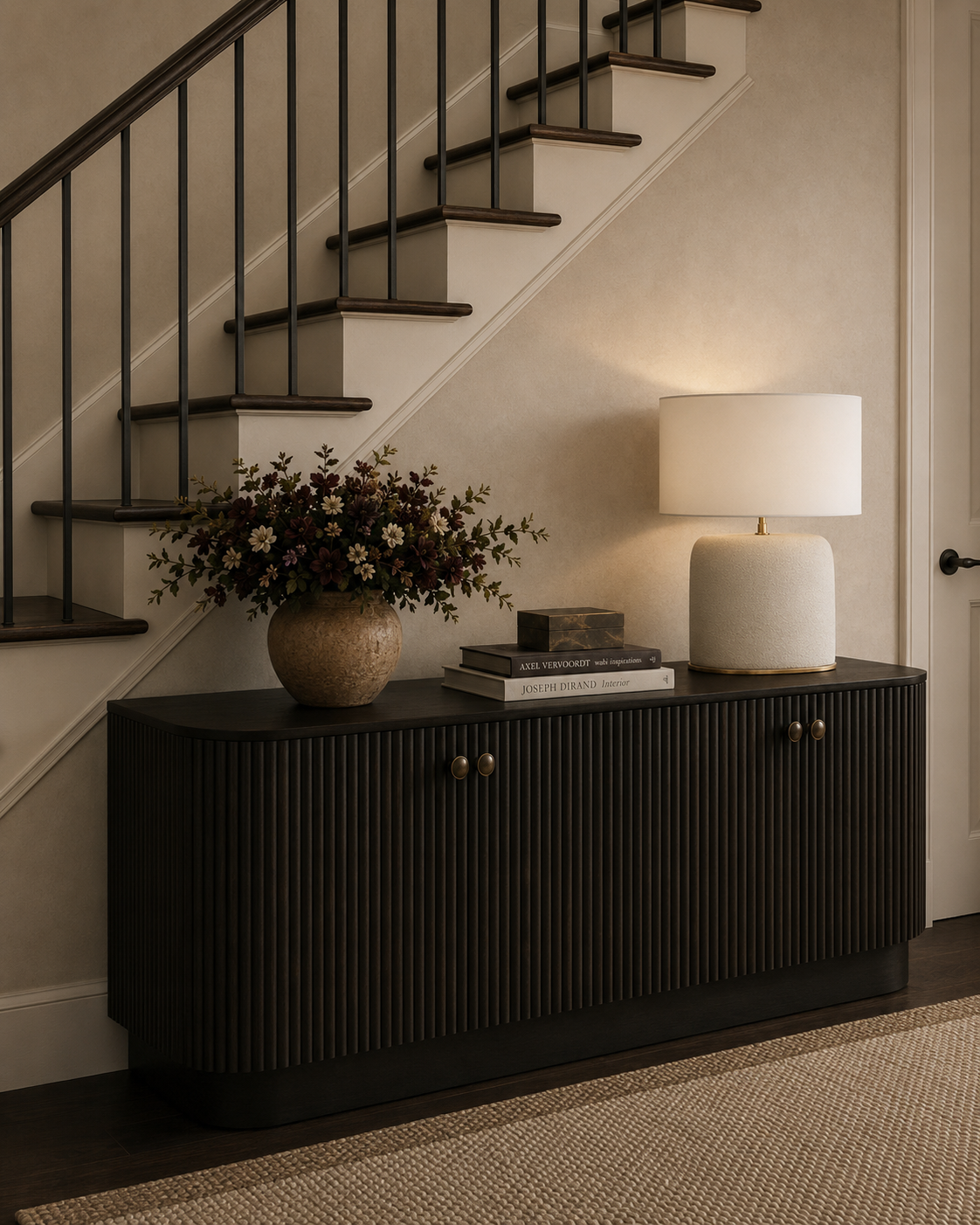A black sideboard with vertical grooves, topped with a bouquet of flowers in a beige vase, several books, and a white table lamp with a large shade, positioned under a staircase with wooden steps and metal railing.