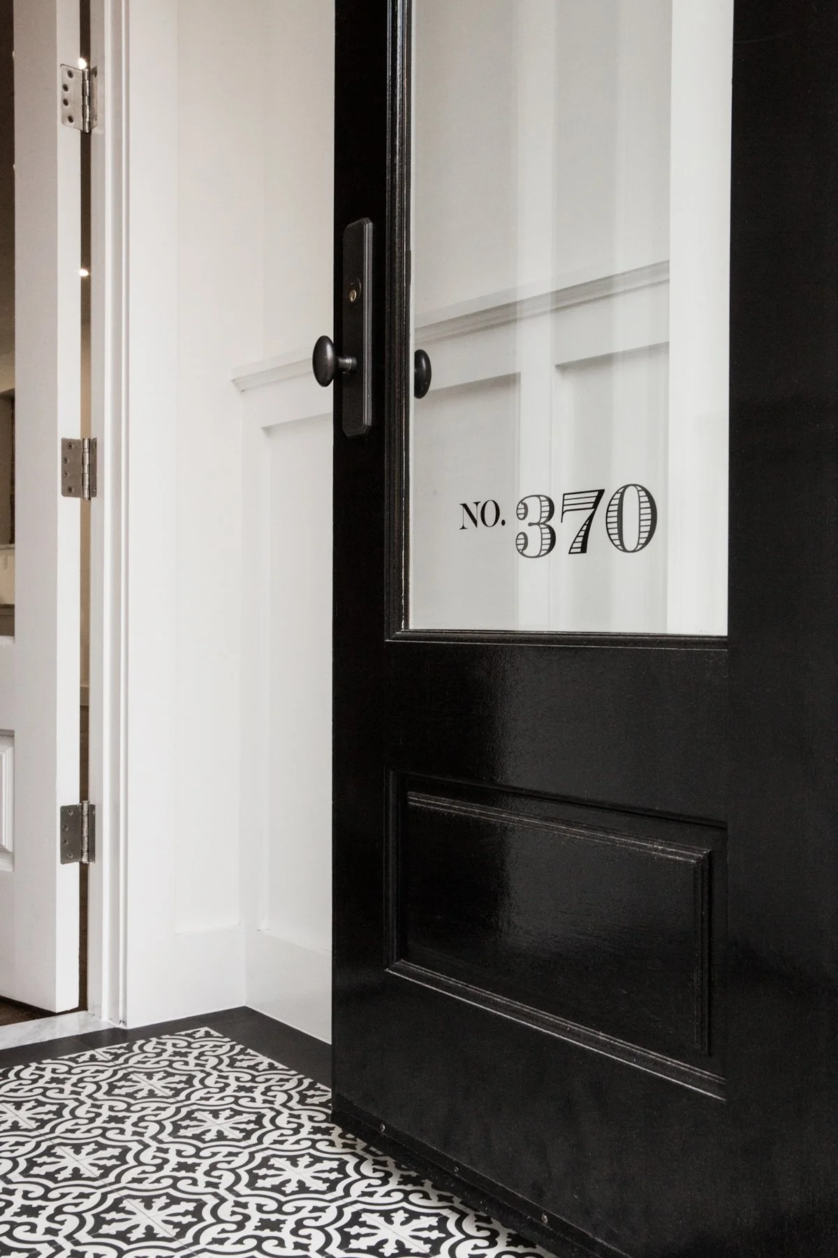 Close-up of a black front door with a glass pane in a restored deco doorway, next to a white interior door, with patterned black-and-white tiles on the floor. The doorway appears luxurious and modern with old world details and superior craftsmanship.