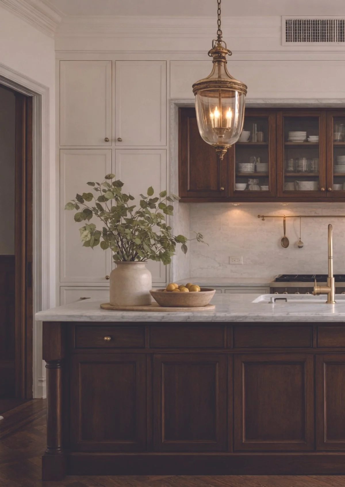 A kitchen with a dark wood island and white marble countertop, a beige vase with green leaves, a wooden bowl with lemons, gold hardware, and a vintage hanging light fixture.