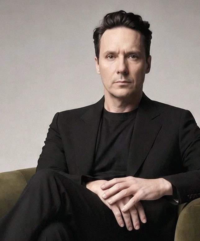 A man with dark hair and light skin, dressed in a Evan Kinori  suit and black shirt, sitting on a greenish-brown chair with one leg crossed over the other, looking directly at the camera with a serious expression.