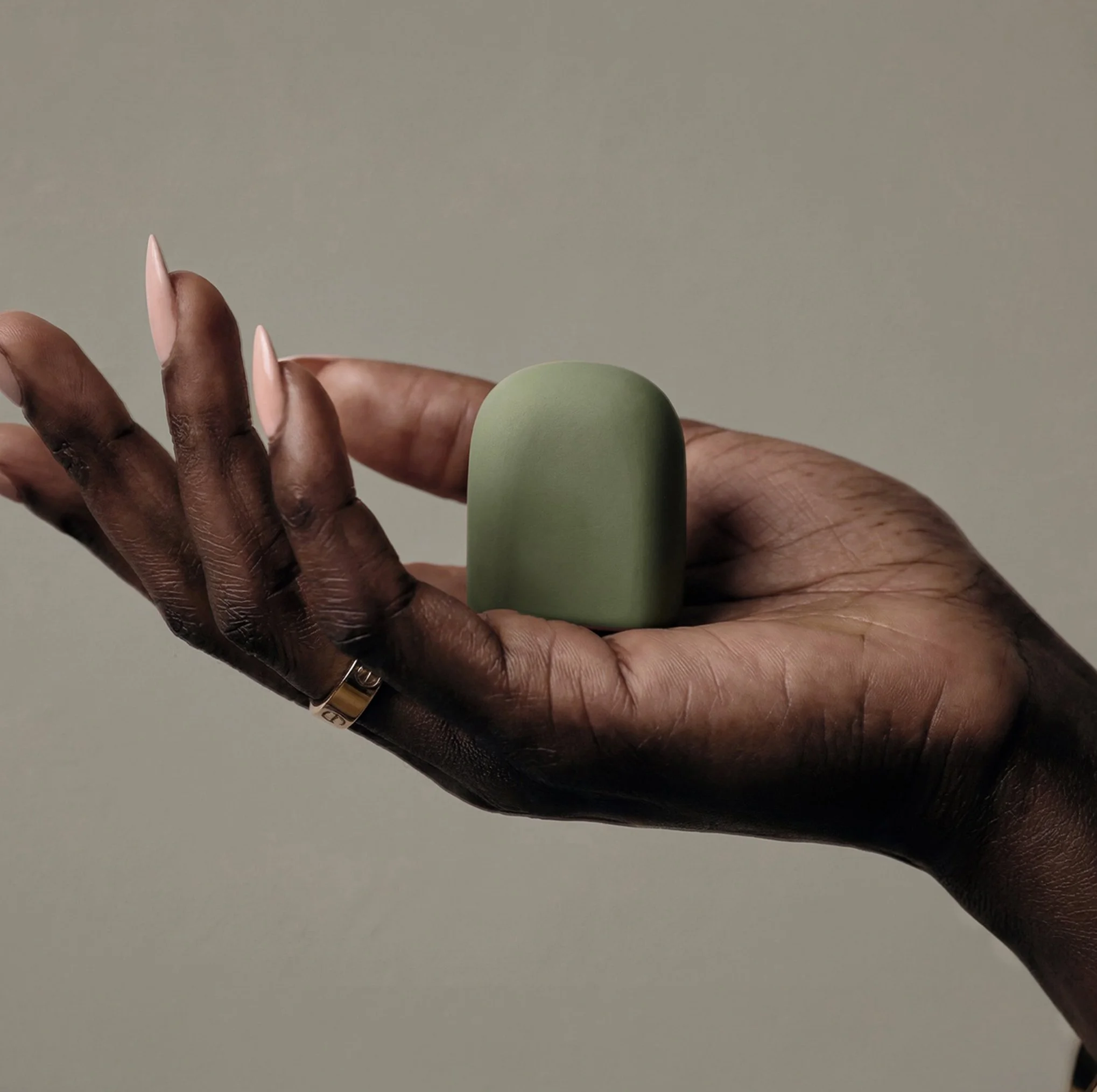 A hand with dark skin and long, pink, almond-shaped nails holding a small, round, modern drab green Omnipod Pod Insulin Cover.