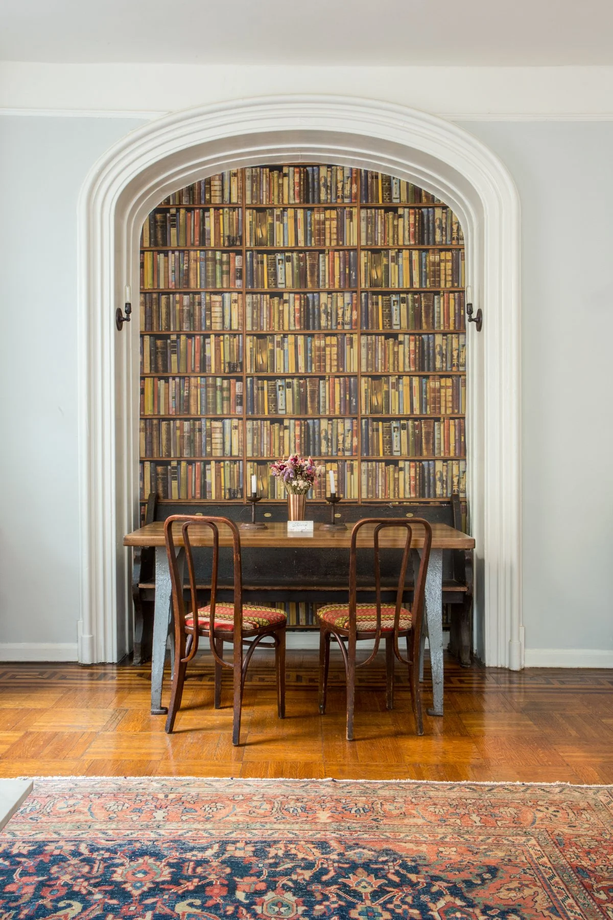 A small dining nook with a wooden table, two chairs with patterned cushions, a flower arrangement, and a bookshelf wallpapered wall.  The space appears to be a design solution within a sophisticated home, where an original design was modernized.