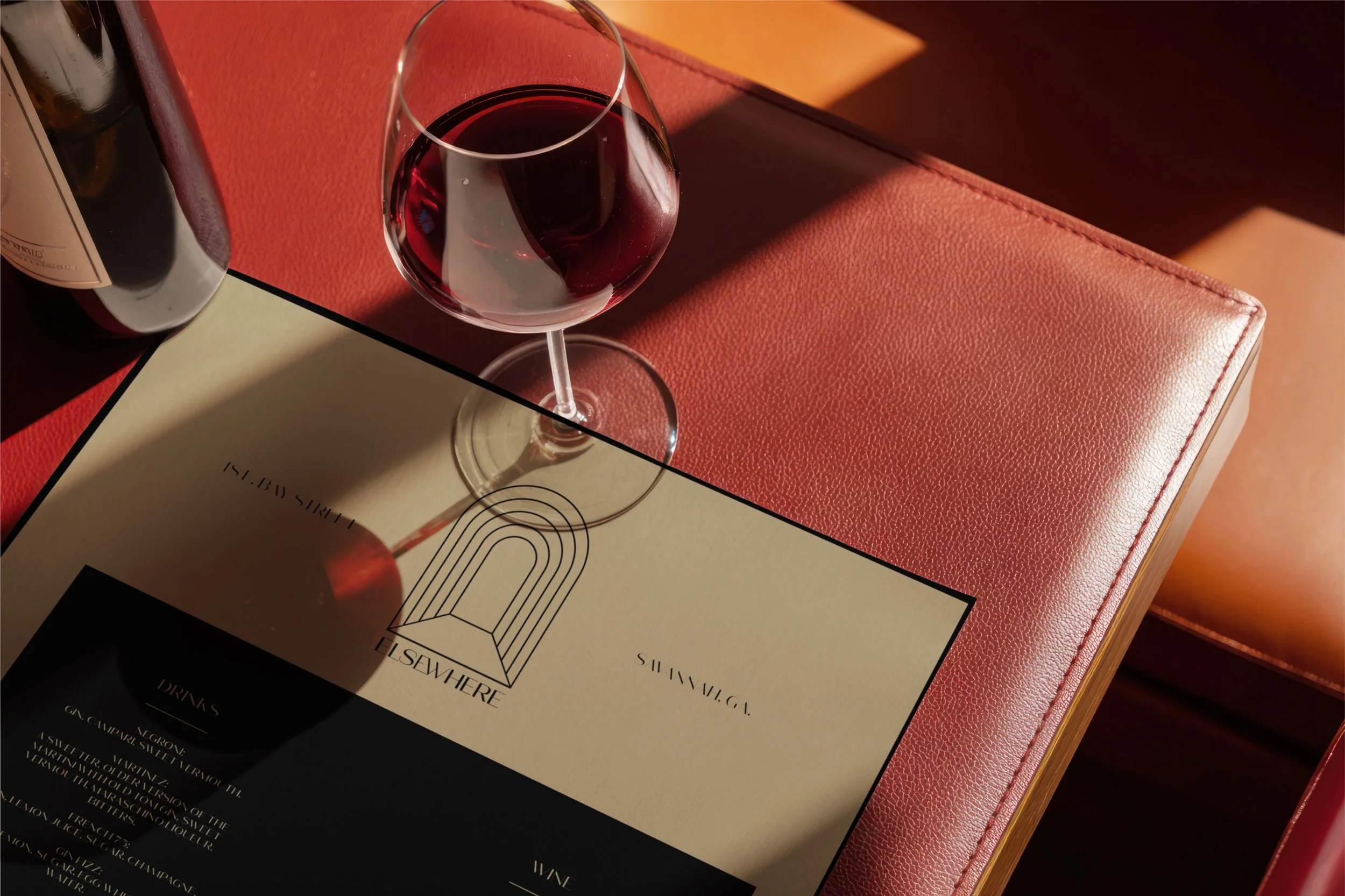 A glass of red wine on a table next to a wine bottle and a menu behind it. The table has a textured pink surface and warm lighting.