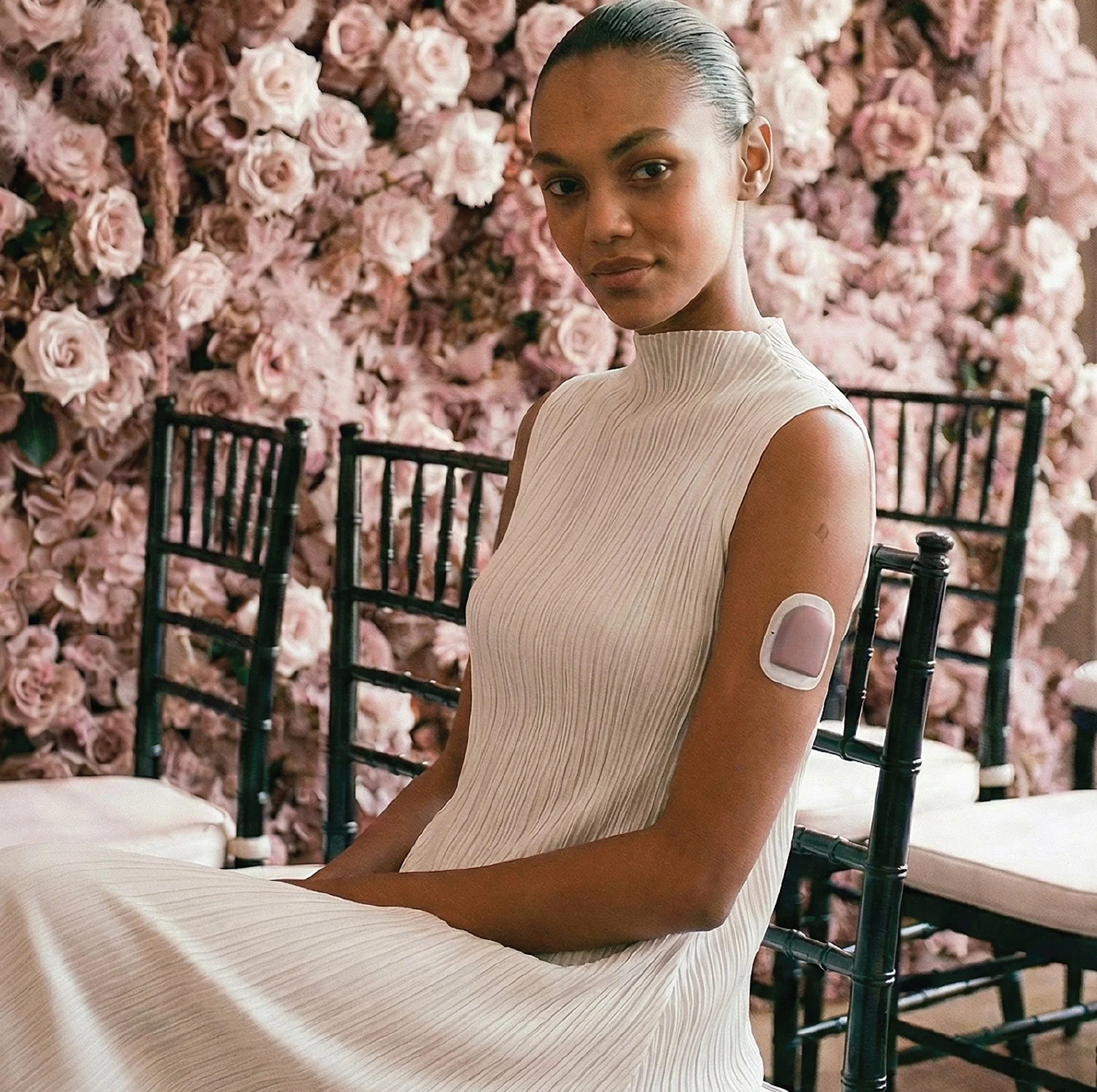 A woman with slicked back hair and natural makeup sitting on a black chair in front of a wall of pink roses, wearing a sleeveless cream-colored dress. She has a blush colored Omnipod Pump accessory on her arm by a brand called Podern.