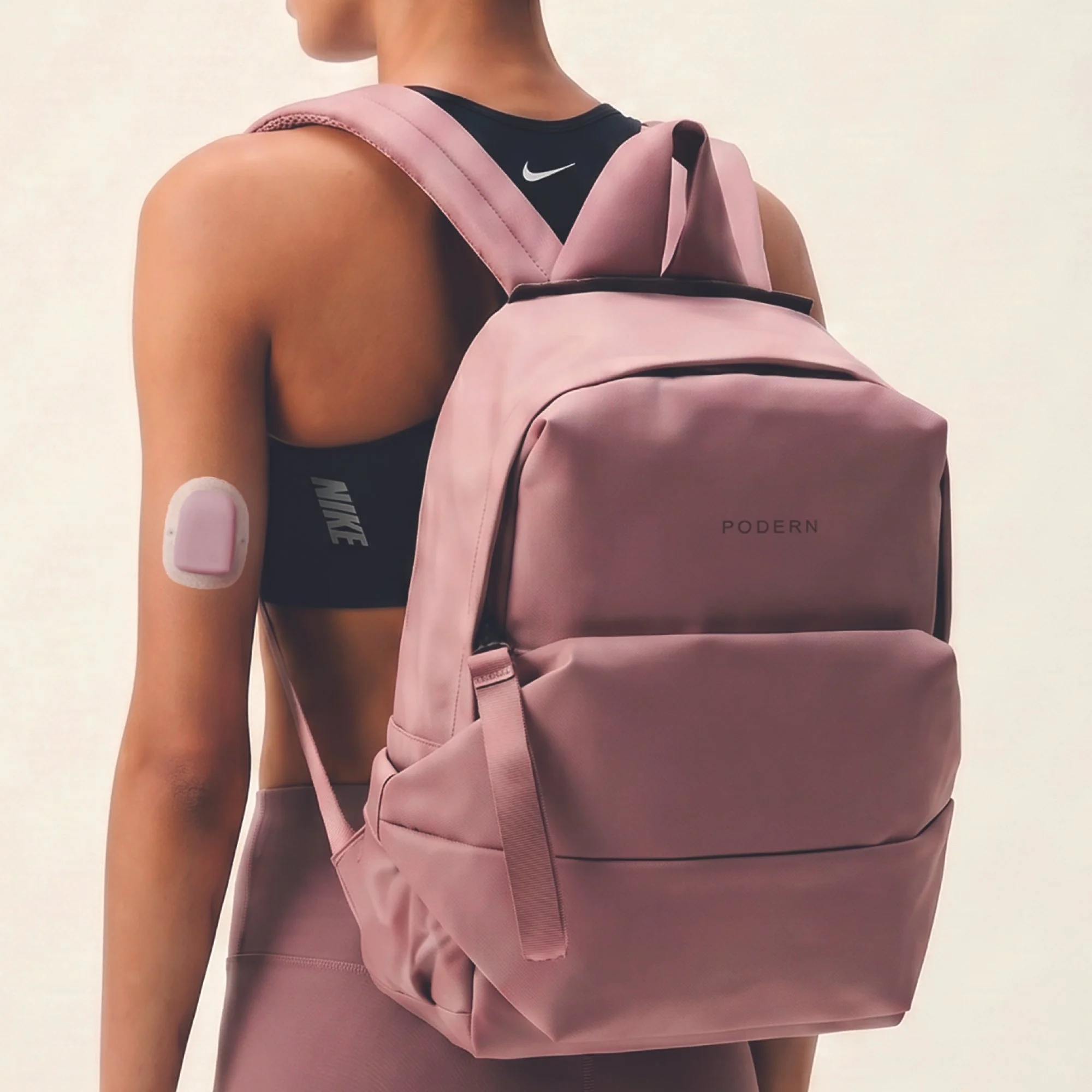 A person with tanned skin wearing a black Nike sports bra and a pink PODERN backpack, carrying a pink purse, against a plain white background.