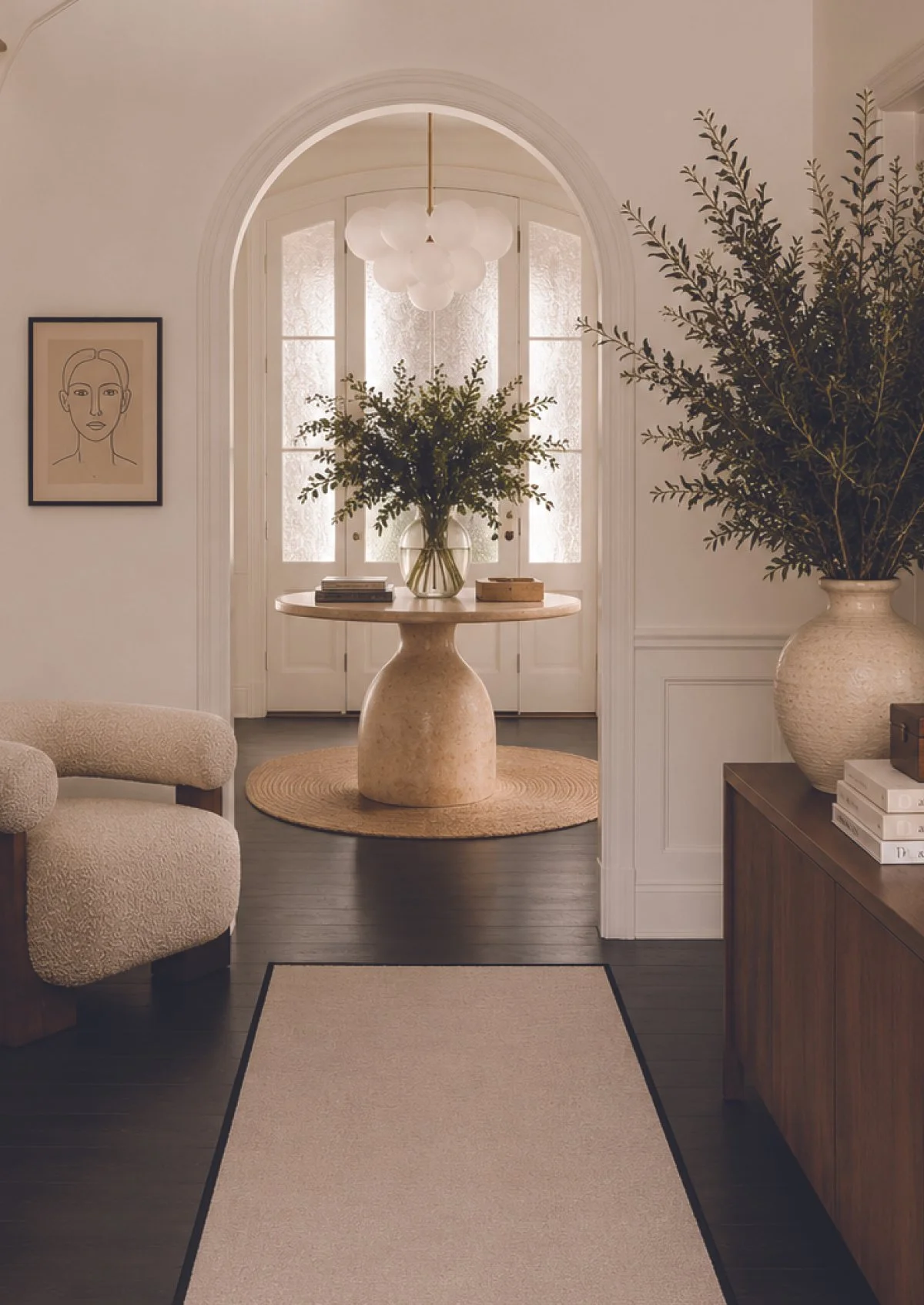 A living room with a view of a white front door with glass panels, a round beige rug, a beige textured armchair, a wooden console table, large potted plants, a framed line art portrait on the wall, and a modern chandelier.