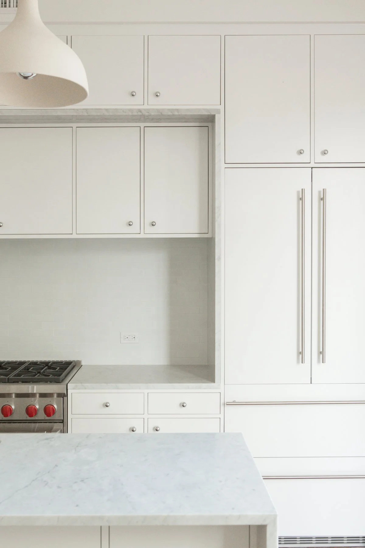 White kitchen with built-in cabinets, marble countertops, a stove with red control knobs, and a large double-door refrigerator.  The use of light and design materials appears very modern and warm, with clean lines you see in Architectural Design.