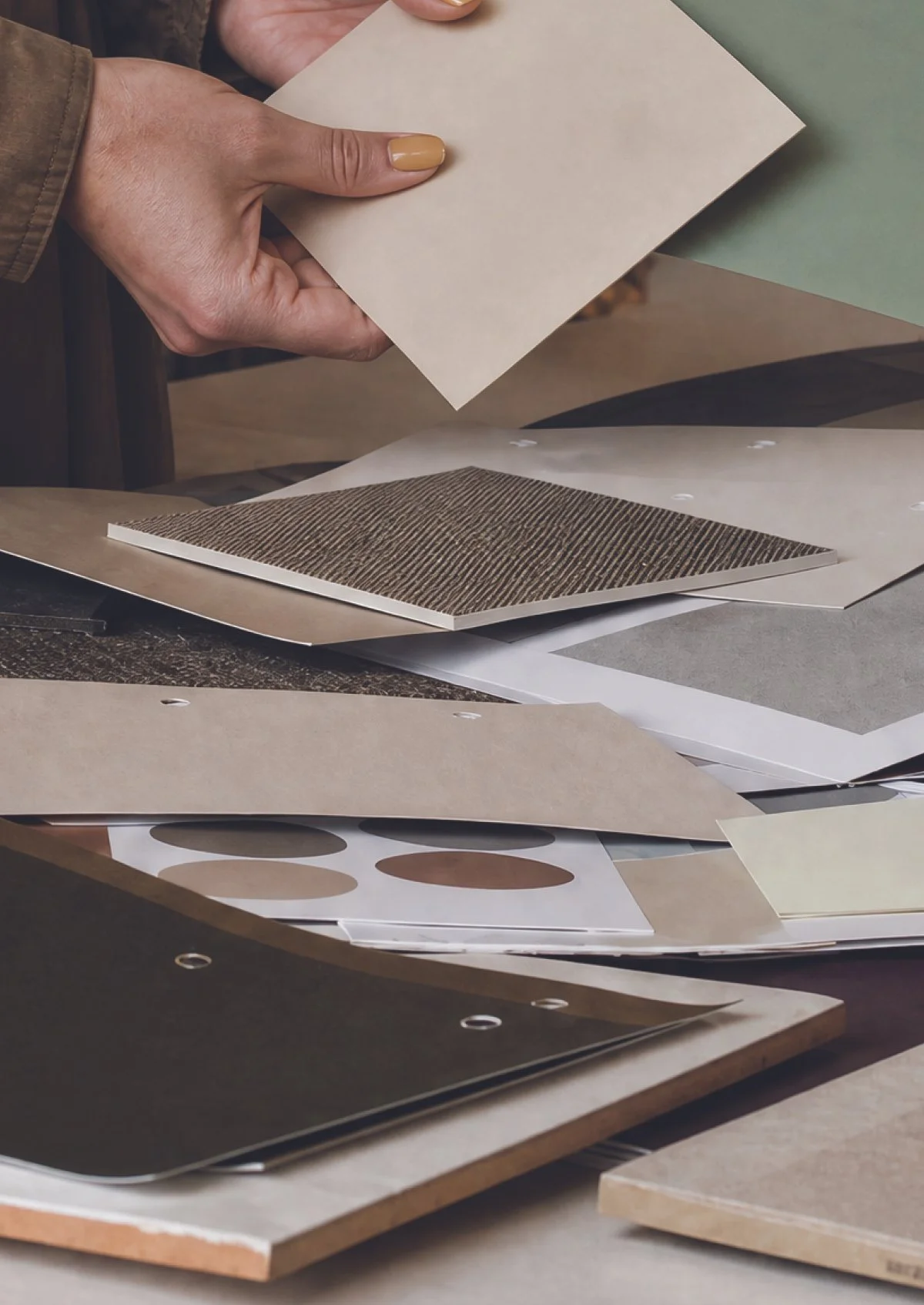 Hands holding a sheet of beige paper over a table with various sample materials and color swatches.