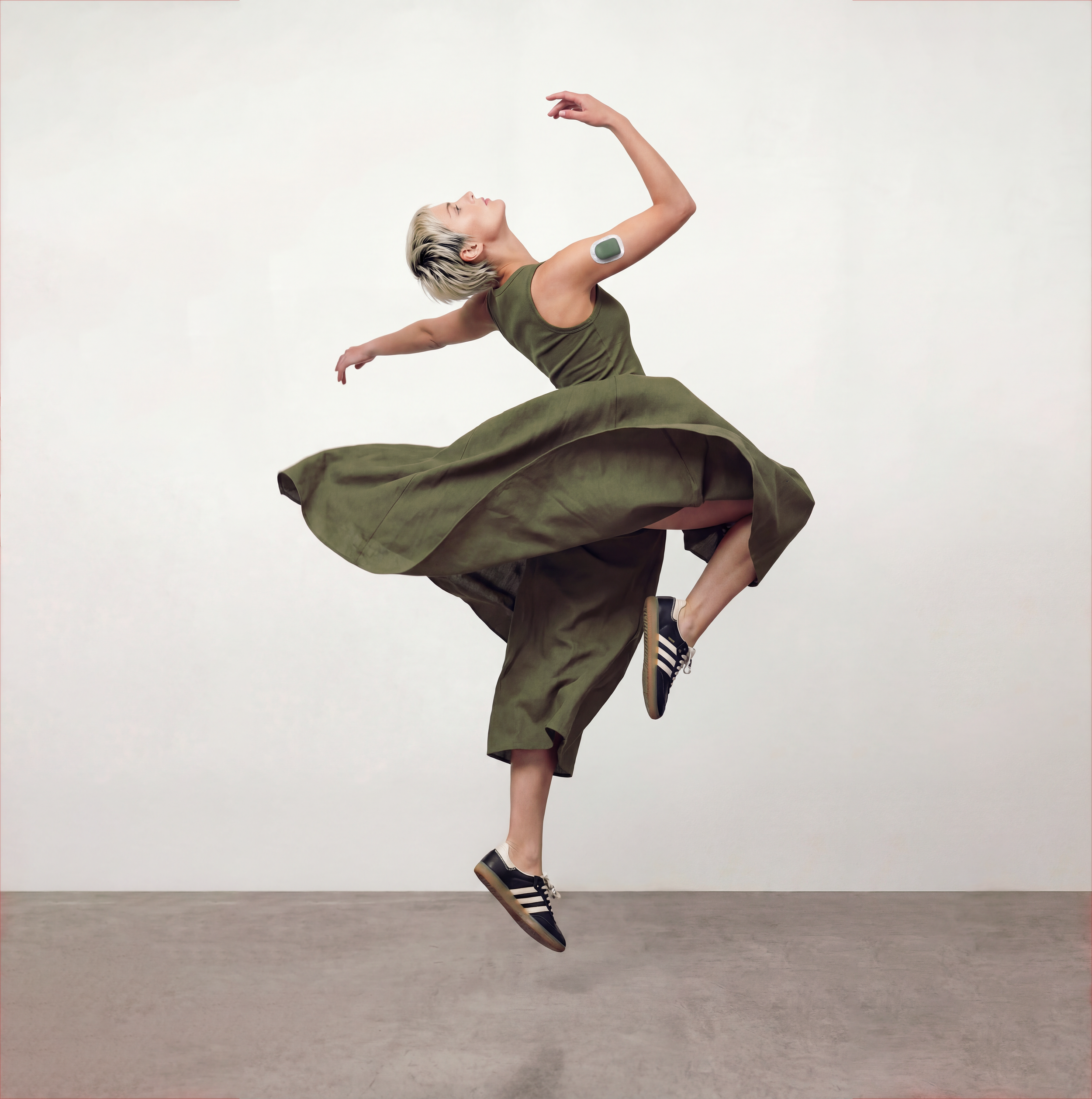A woman with short platinum blonde hair in a green sleeveless outfit is balancing on one leg and stretching her opposite arm upward. She is wearing Wales Bonner Adidas with a Podern Omnipod Cover on her insulin pump.