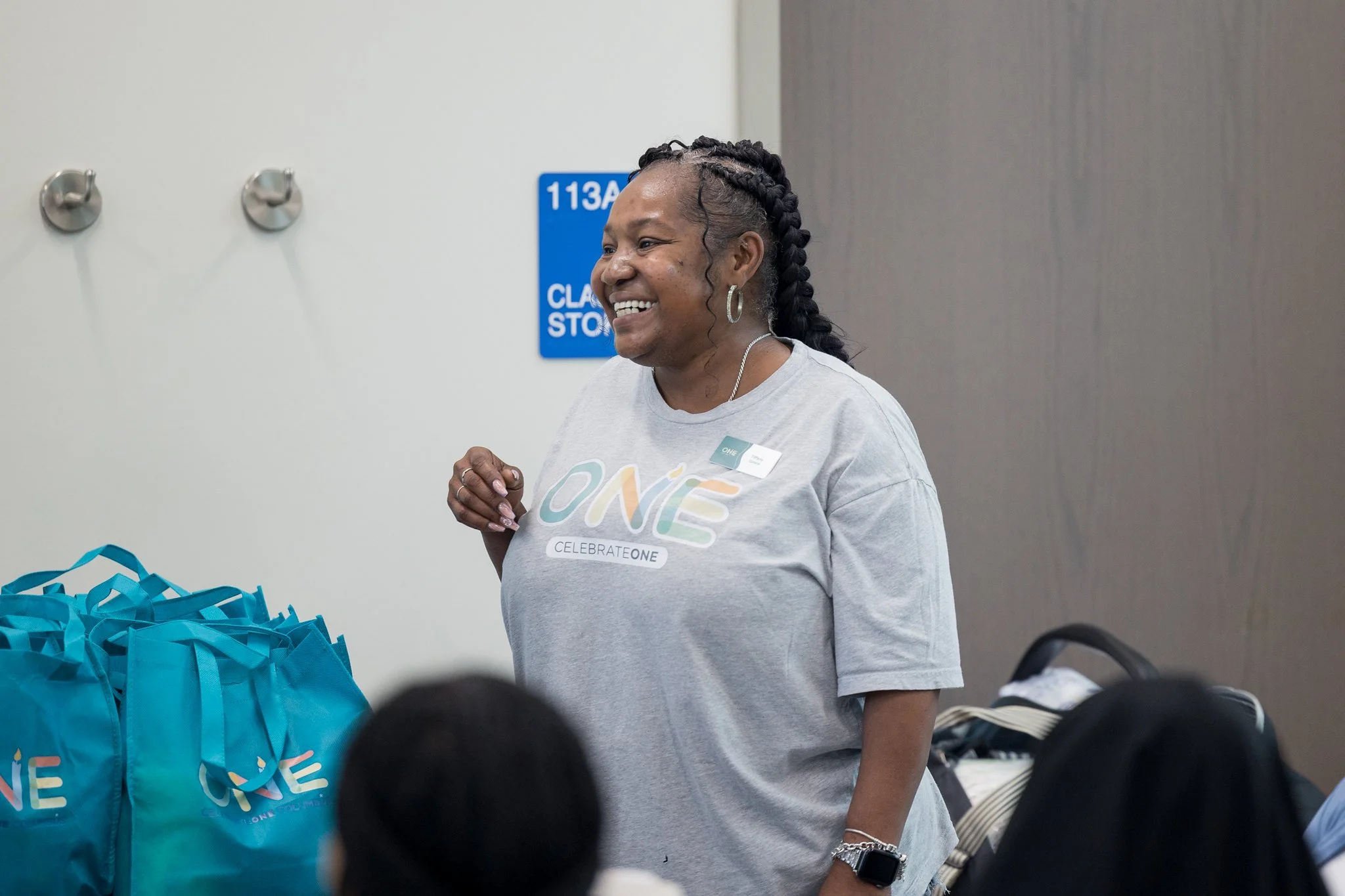 A smiling woman wearing a grey shirt with 'ONE' logo and a name badge, standing in front of blue bags with the same logo, at an indoor event.