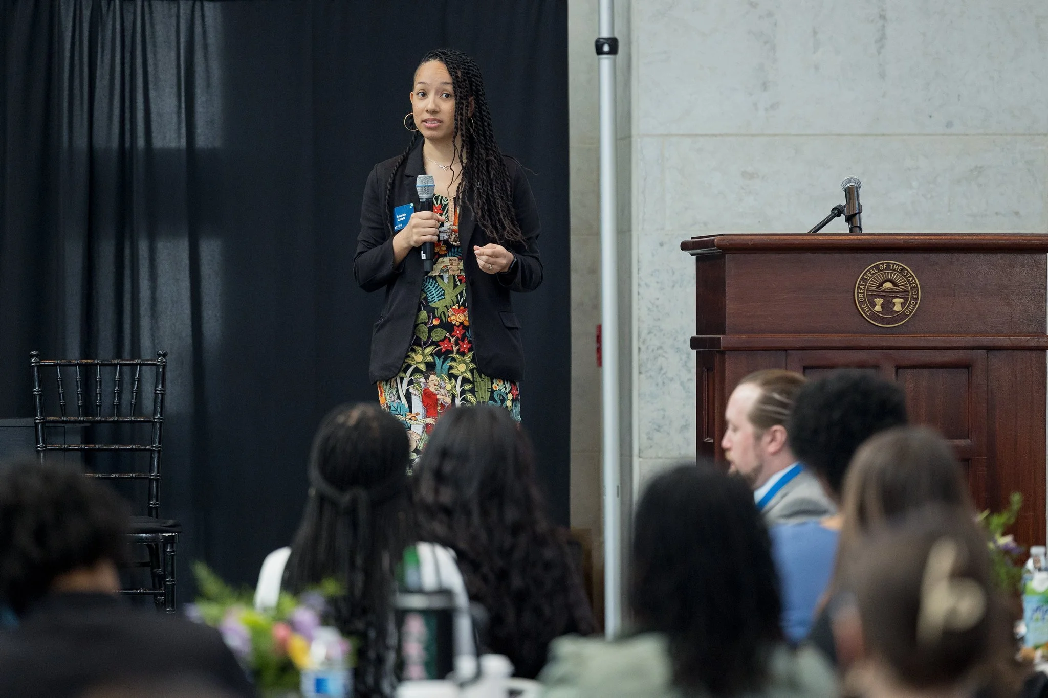 A woman with long braids wearing a colorful dress and black blazer speaks into a microphone on a stage at a formal event, with an audience seated in front of her.