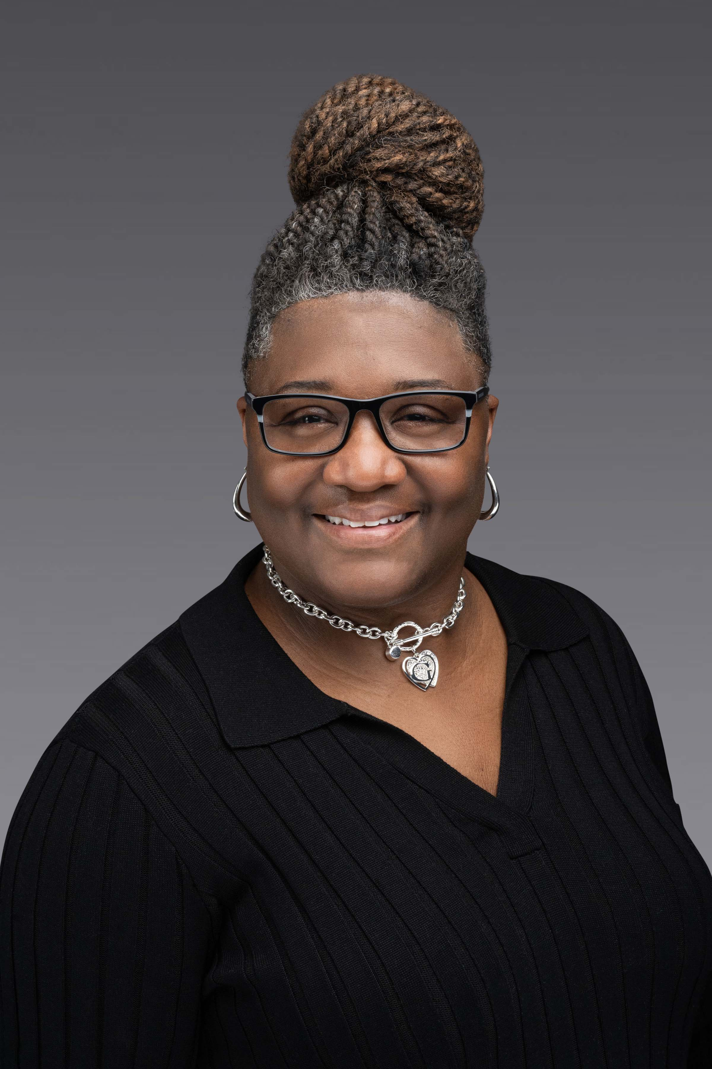 Portrait of a smiling a woman wearing glasses, hoop earrings, and a heart-shaped pendant necklace against a gray background.