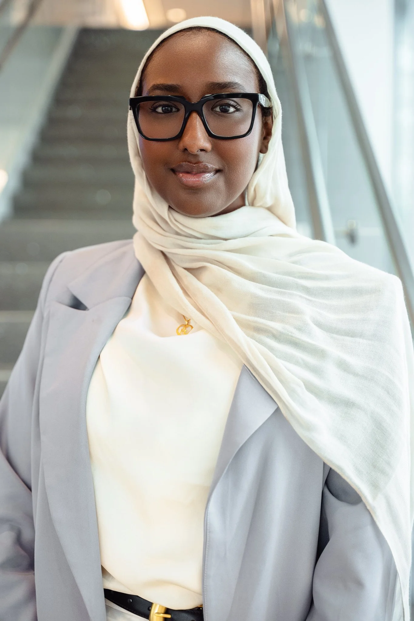 Professional woman wearing glasses, hijab, light gray blazer, and a white shirt, standing in a modern building with stairs in the background.