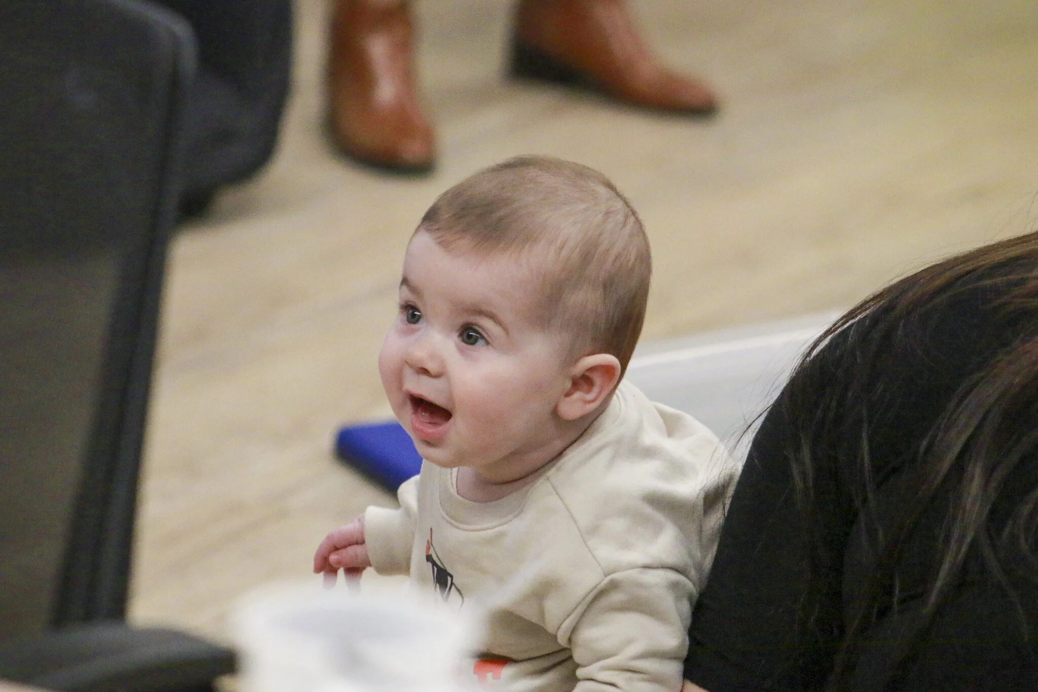 A baby with brown hair and blue eyes looking amazed, seated on the floor.
