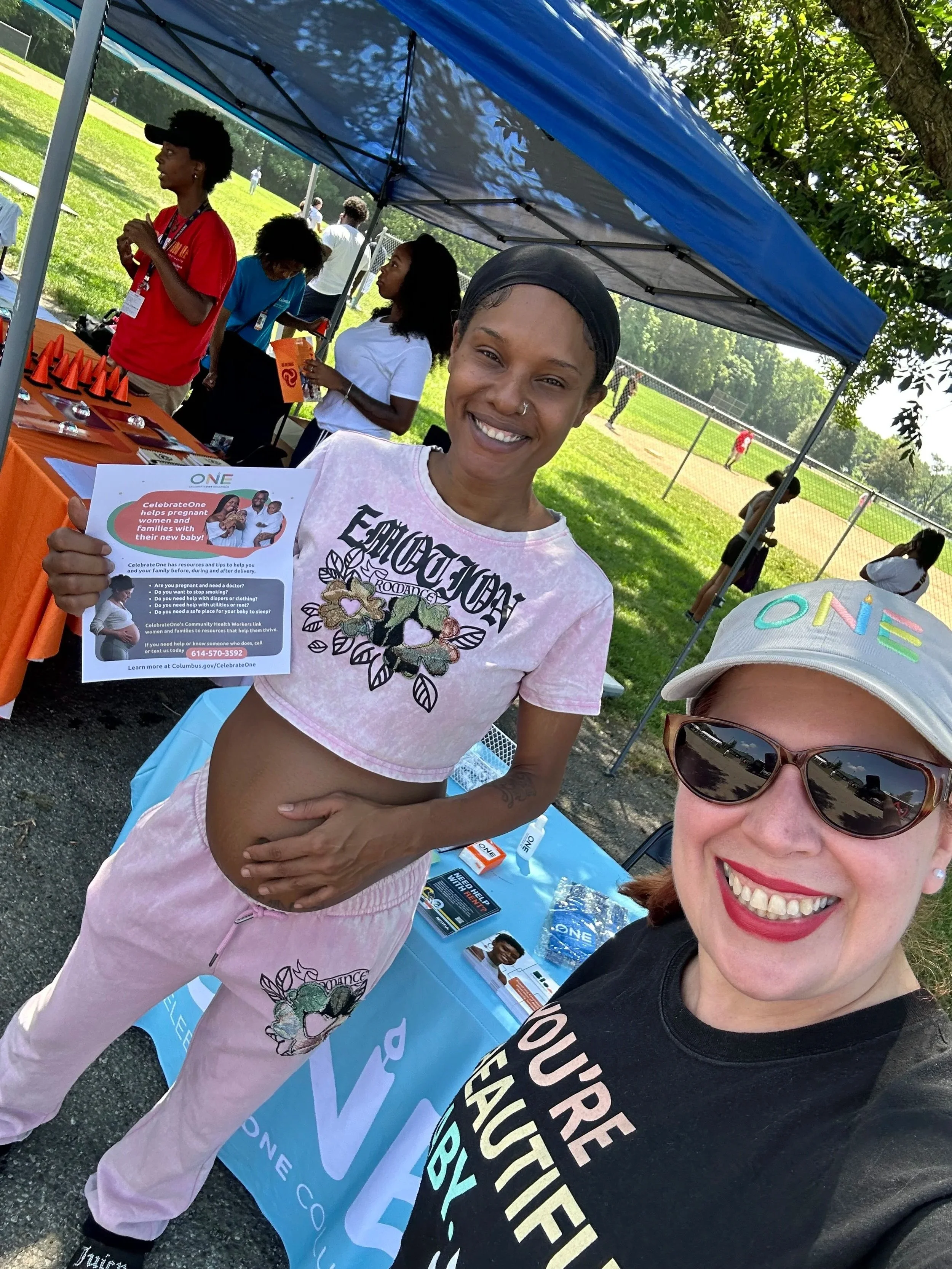 Two women smiling at an outdoor health awareness event. One woman is pregnant, wearing a pink and white outfit, holding a flyer about a health program. The other woman is wearing sunglasses and a cap with colorful writing, and a black T-shirt. They are standing under a blue canopy, with a table displaying pamphlets and brochures, and a baseball field in the background.