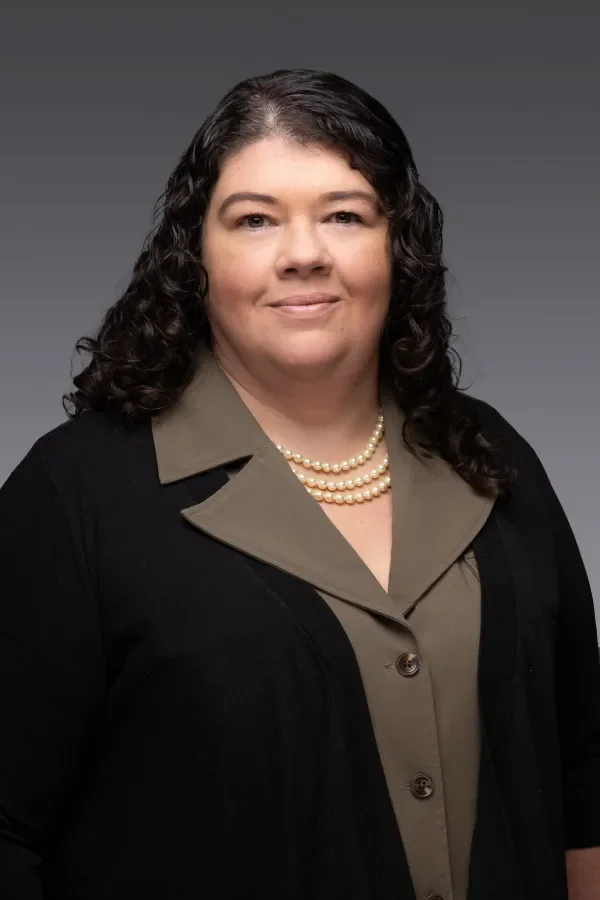 A woman with dark, curly hair wearing a khaki blouse, a black cardigan, and a pearl necklace, standing against a gray background.