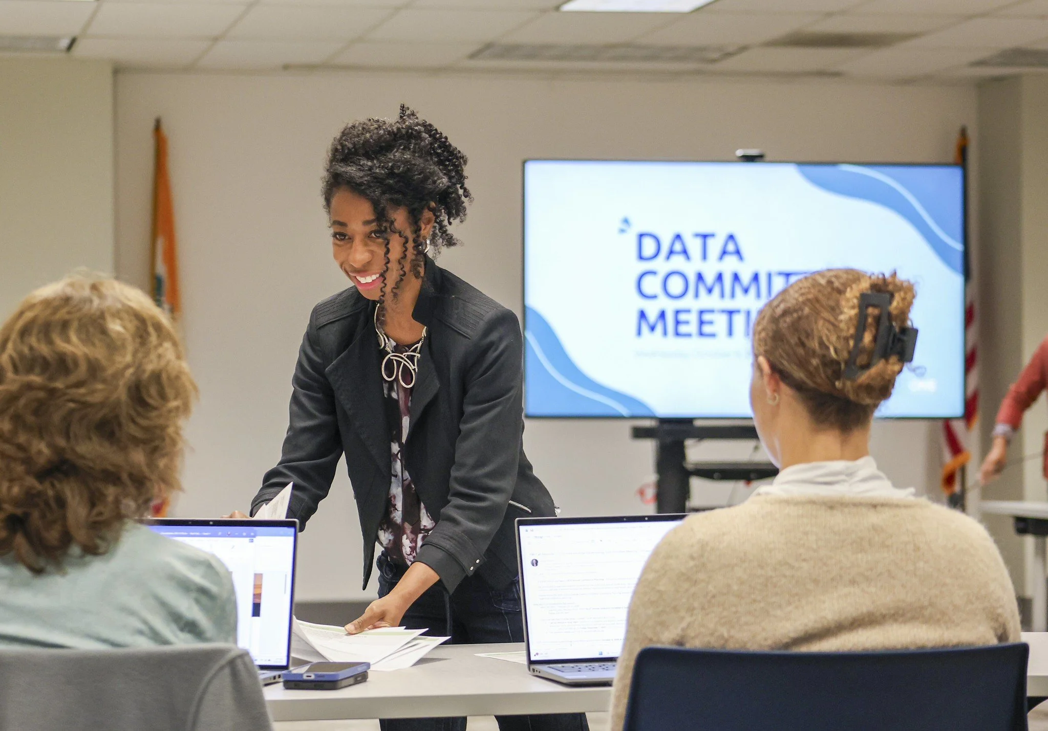 CelebrateOne Director, Danielle Tong, holds a computer in front of a group of people. "Data Committee Meeting" is on the tv screen behind her head