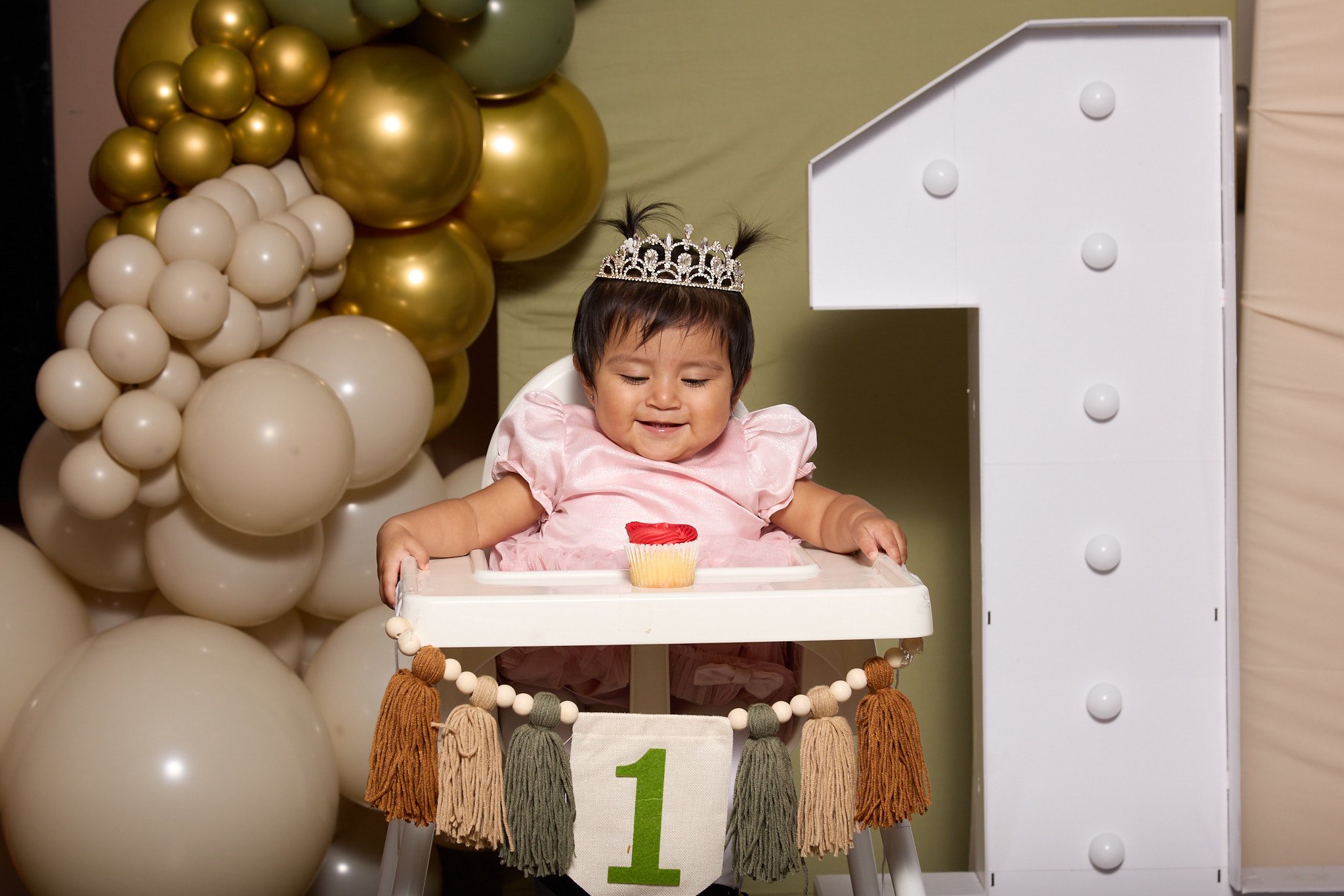 A baby girl in a pink dress with a tiara, sitting in a high chair with a cupcake, celebrating her first birthday. There are white and gold balloons and a large white number 1 decoration in the background.
