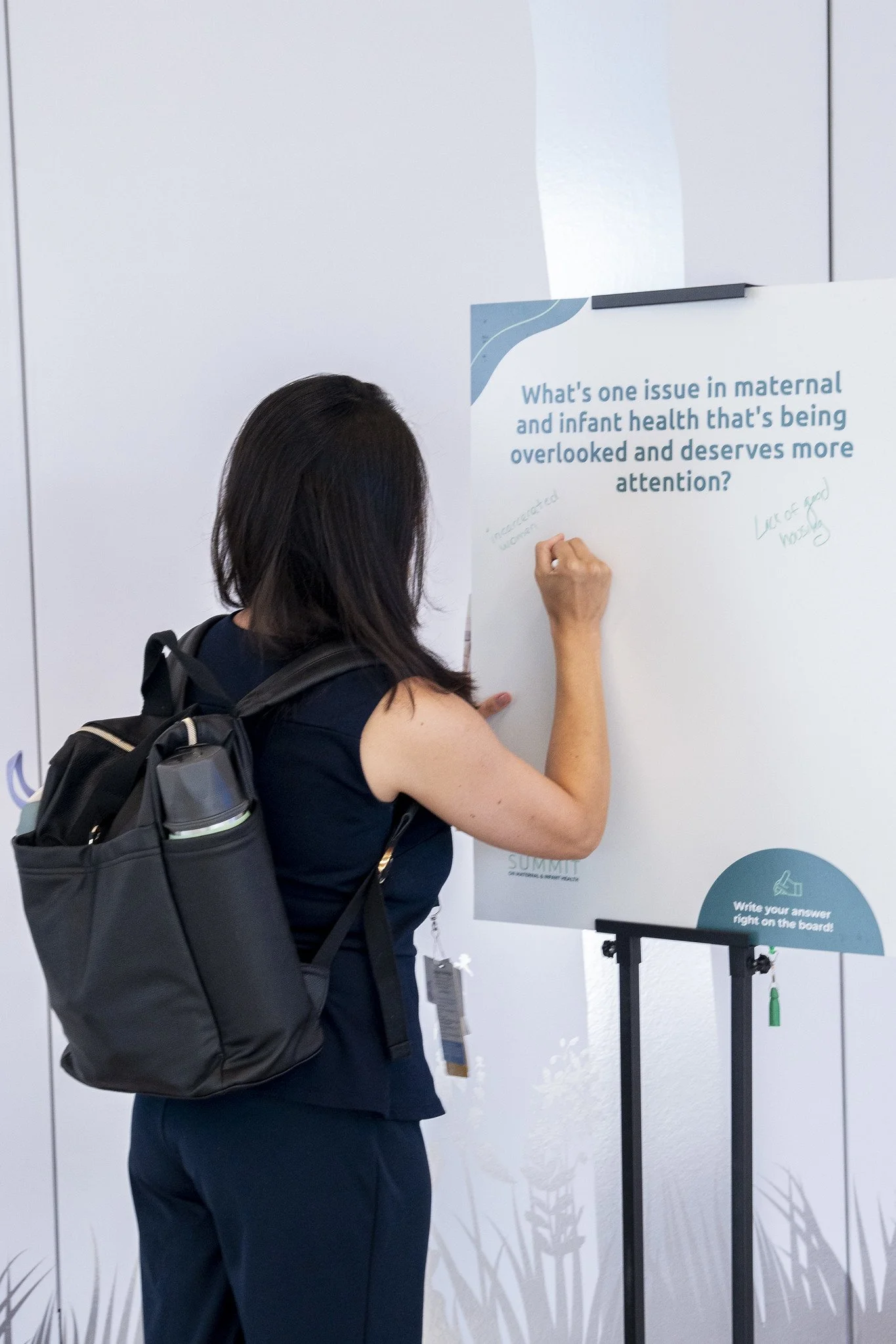 A woman with a backpack writing on a whiteboard at a conference or event.