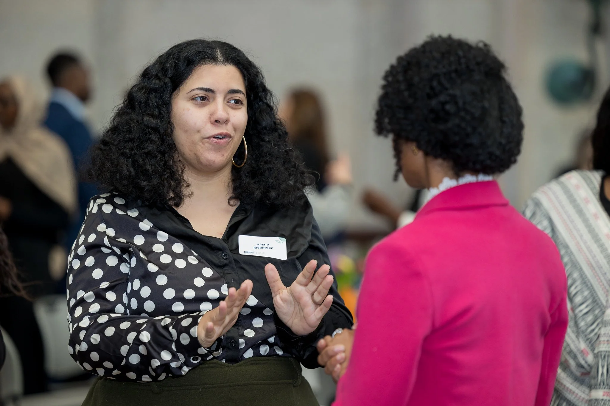 Two women engaging in conversation at a professional event. One woman, with dark curly hair and a black and white polka dot shirt, gestures with her hands. The other woman, with short curly hair and wearing a bright pink blazer, listens attentively. Background shows other attendees mingling.
