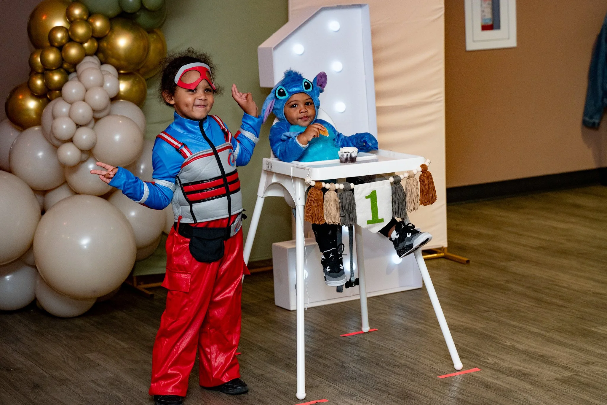 Two children in colorful costumes celebrating a birthday, one dressed as a superhero and the other as Stitch from Lilo & Stitch, sitting in a white high chair decorated with a green number 1, with a backdrop of gold and white balloons.