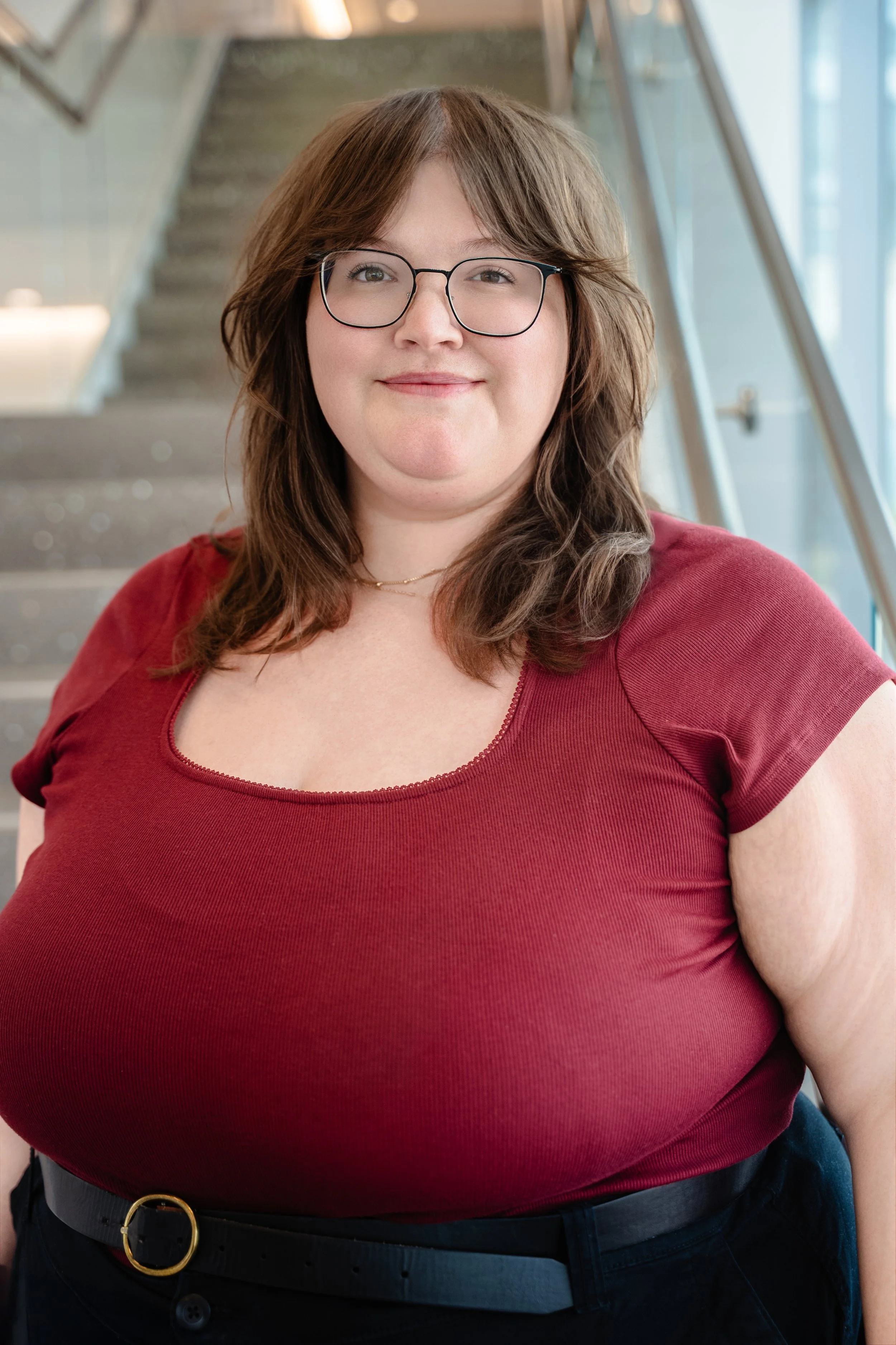 A woman with brown hair, glasses, and a red top standing indoors near a staircase and glass window.