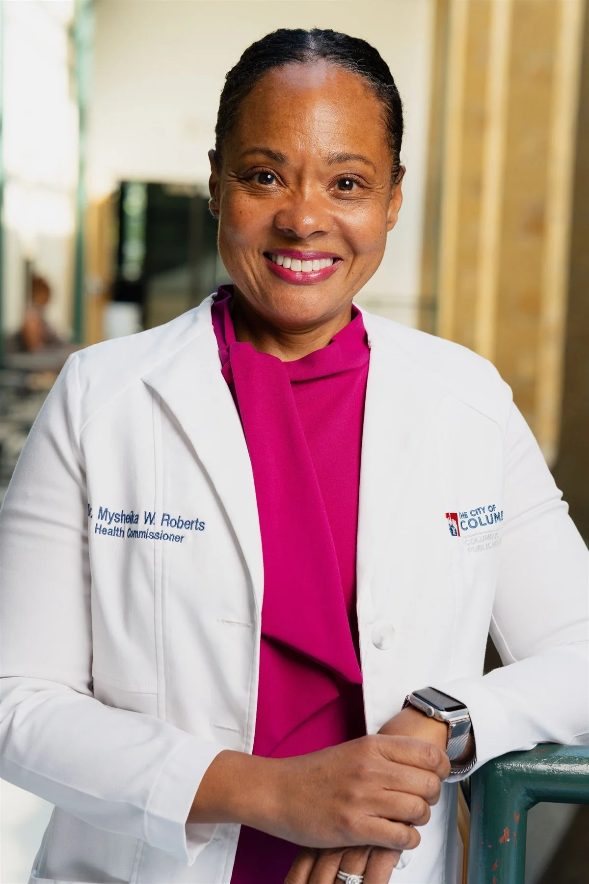 A smiling woman in a white medical coat and magenta top, standing indoors with a blurred background, wearing a smartwatch on her left wrist.