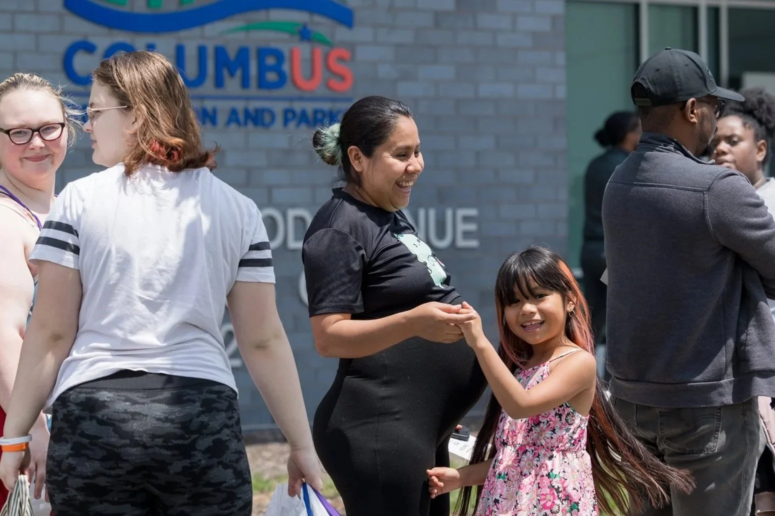 A group of diverse people standing outdoors in front of a building with a sign that reads 'COLUMBUS'. A woman is smiling and holding hands with a young girl who is also smiling, in a casual setting with other people in the background.